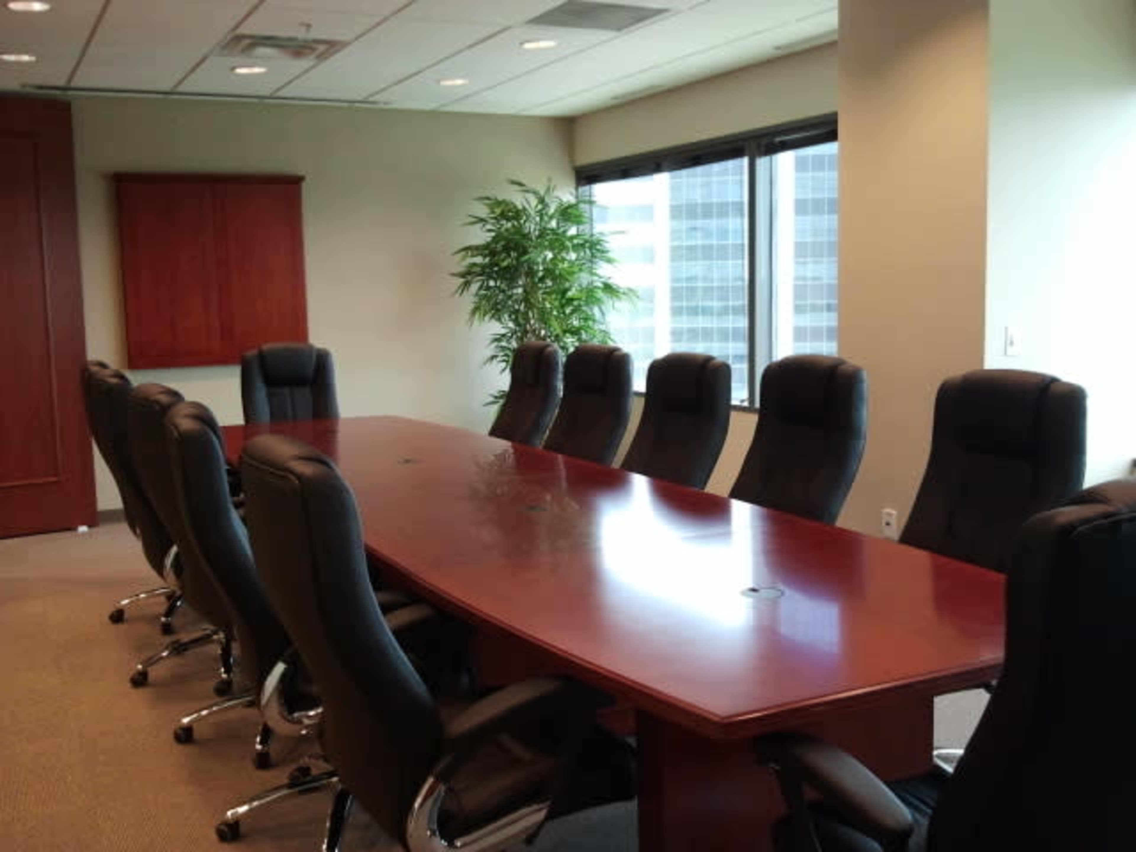A boardroom with a long oval table surrounded by black office chairs and a large window displaying a city view.