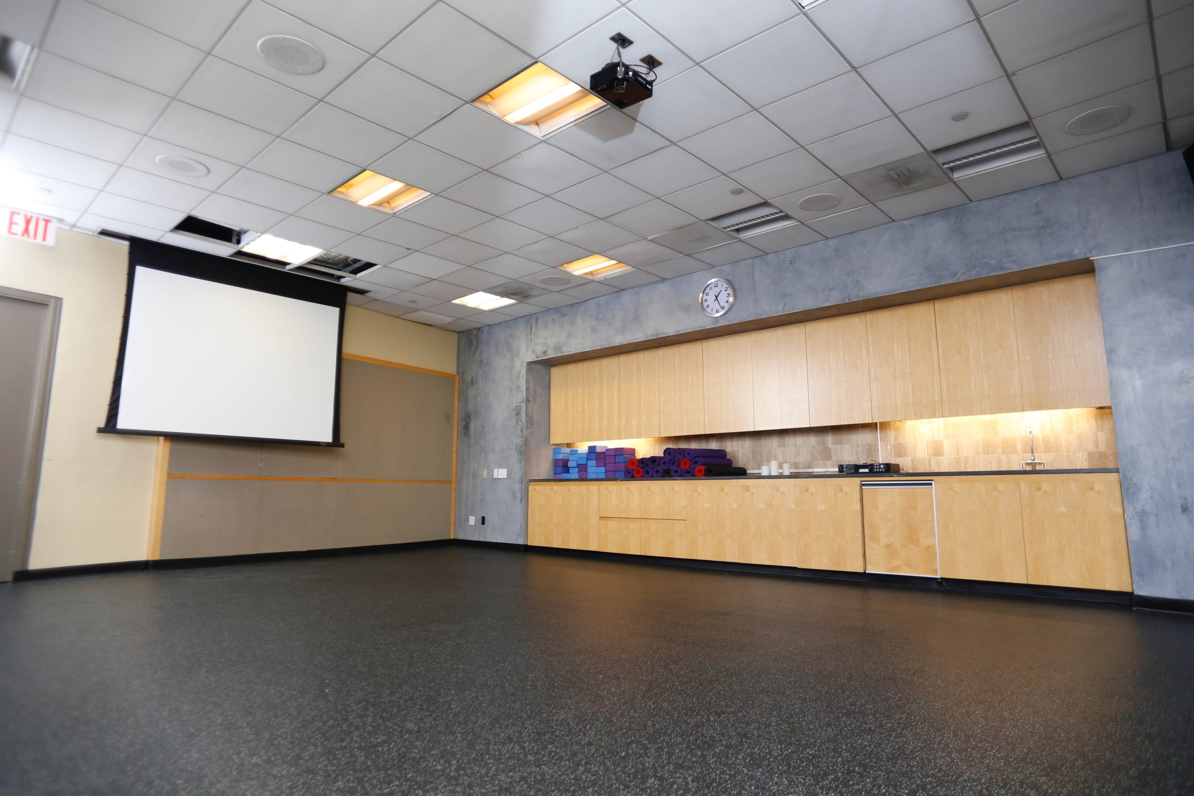 The image shows a spacious room with a simple kitchen area on one wall, a projector mounted on the ceiling, and a large screen pulled down.