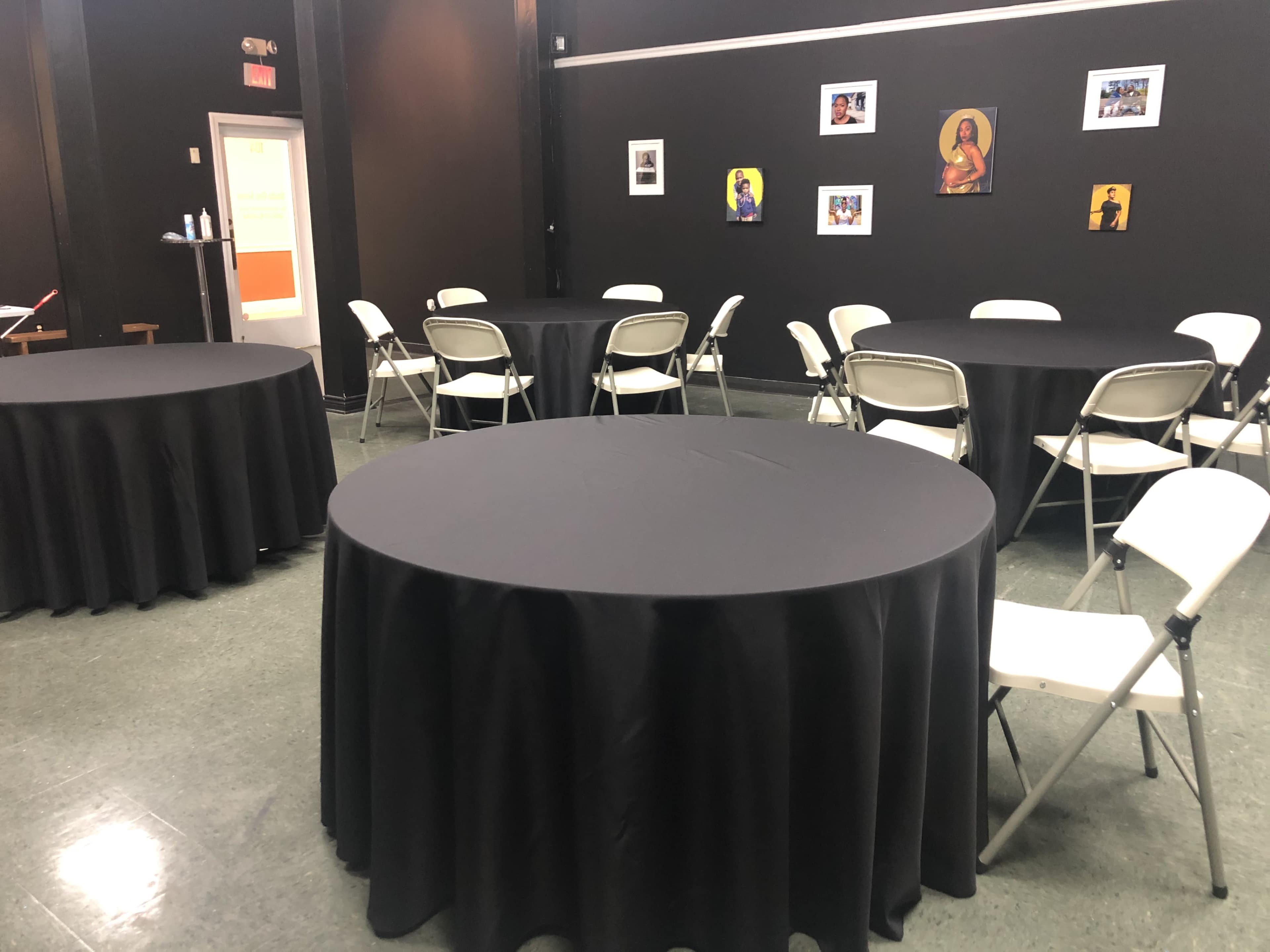A room features several round tables covered with black tablecloths and surrounded by white folding chairs, with framed photographs hanging on the walls.