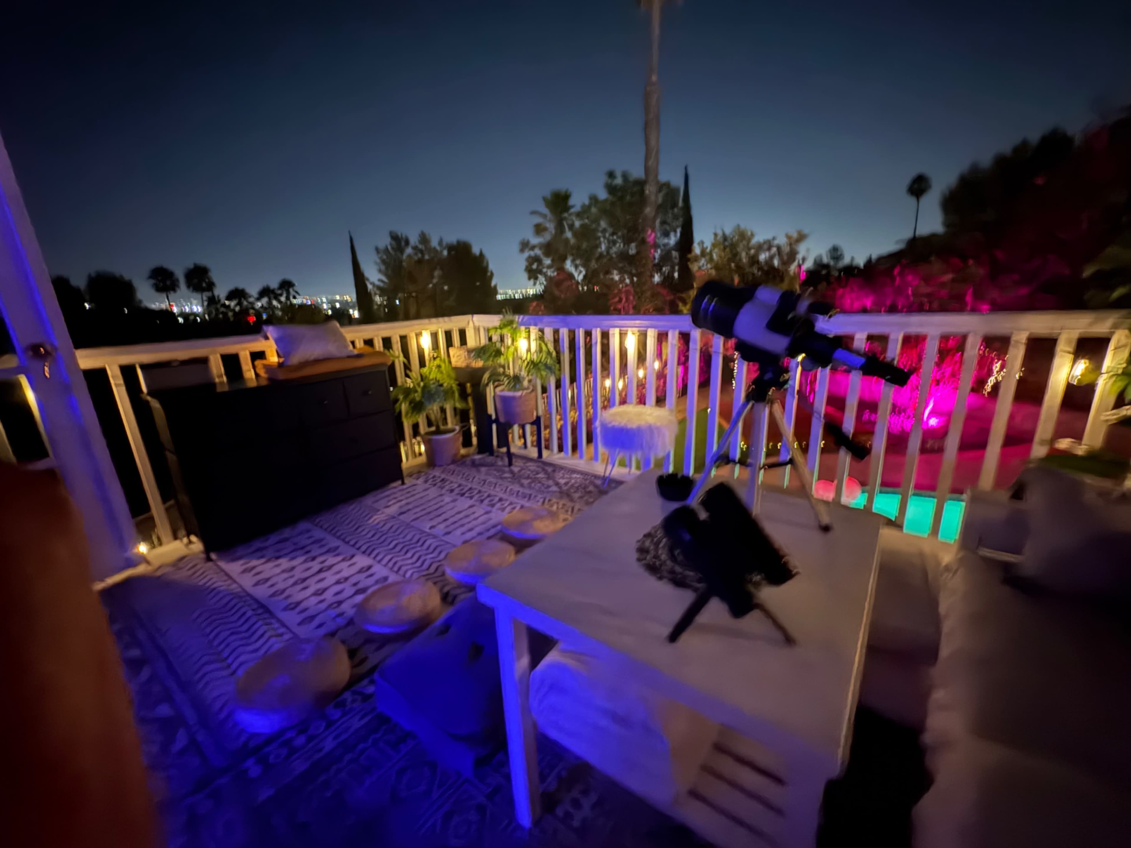 The image depicts a nighttime balcony scene with a telescope set up on a table surrounded by plants and illuminated by colorful lights.