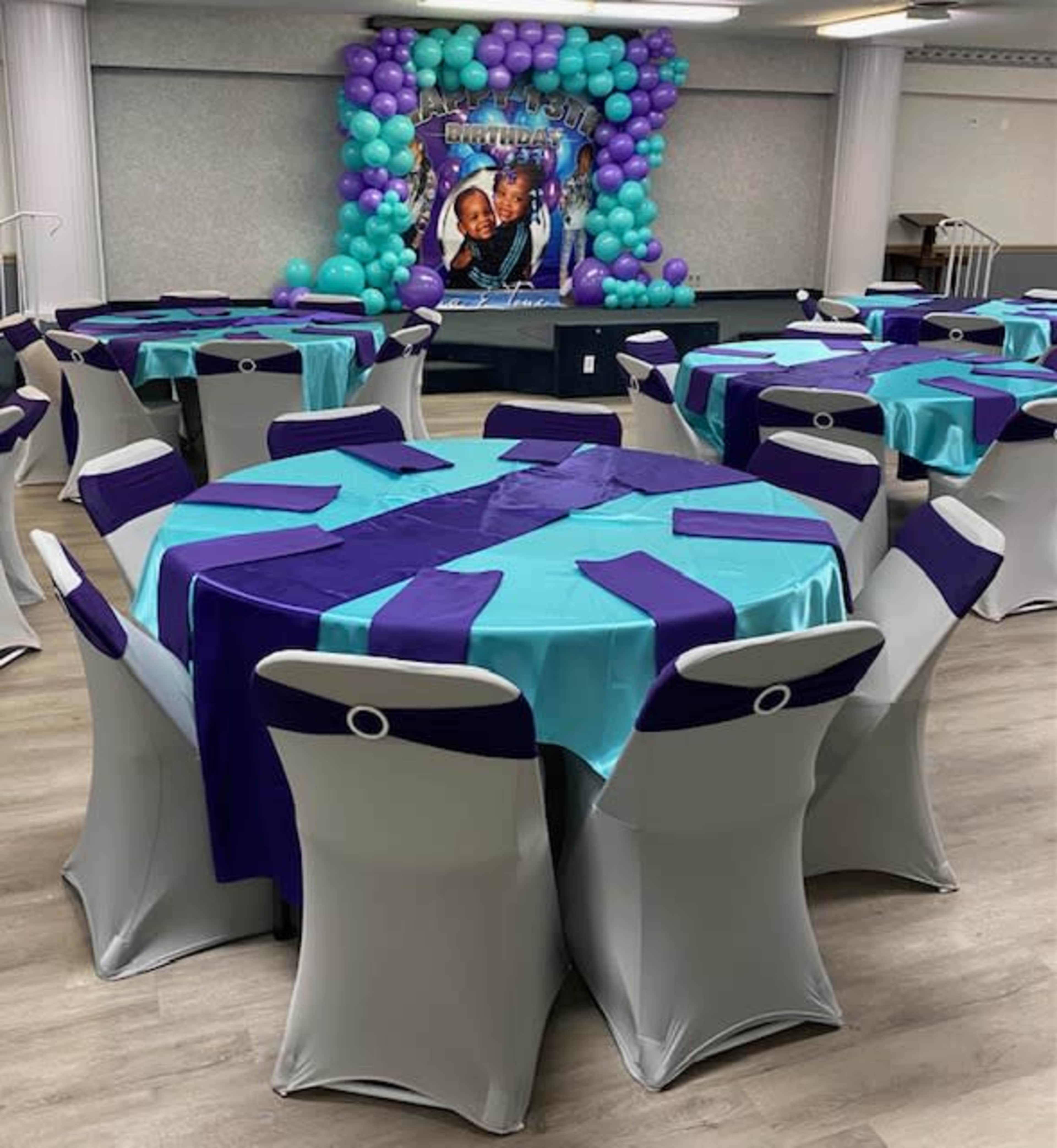 The image shows a banquet hall arranged with several round tables covered in blue and purple tablecloths, surrounded by chairs with matching fabric accents, and a backdrop decorated with balloons.