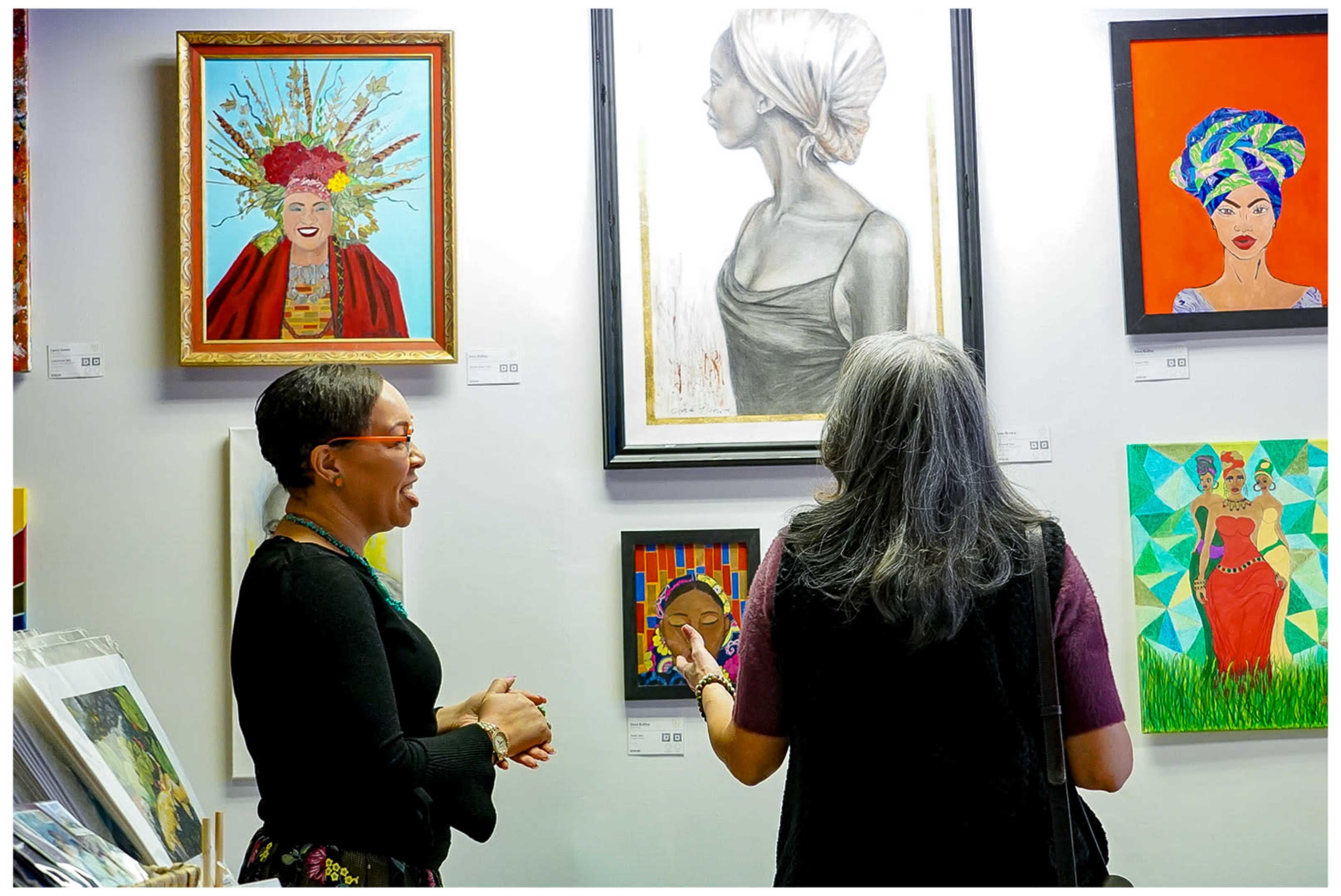 Two women engage in conversation while observing various colorful artworks displayed on a gallery wall.