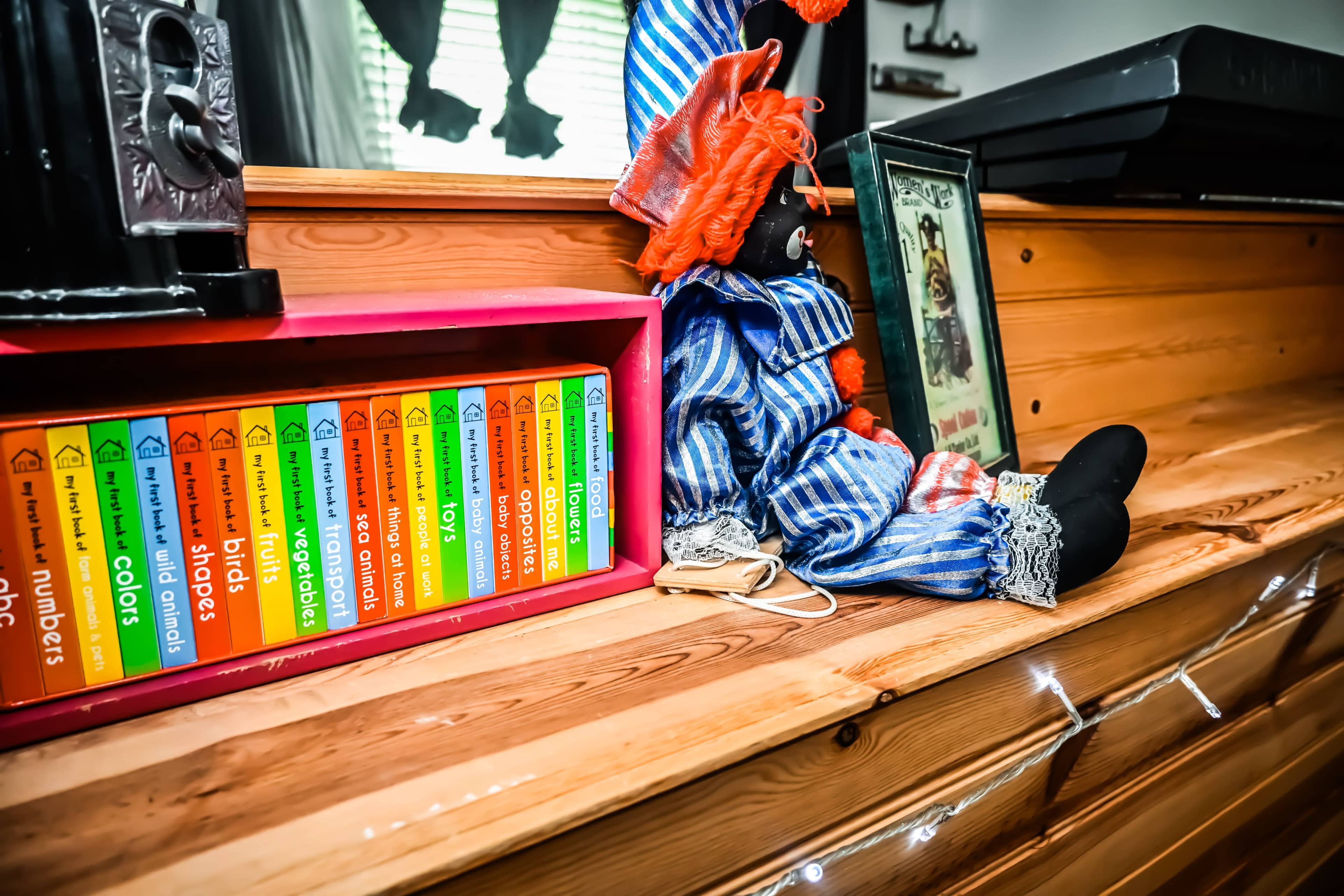 The image shows a wooden shelf displaying colorful books and a clown figure, alongside a framed picture.