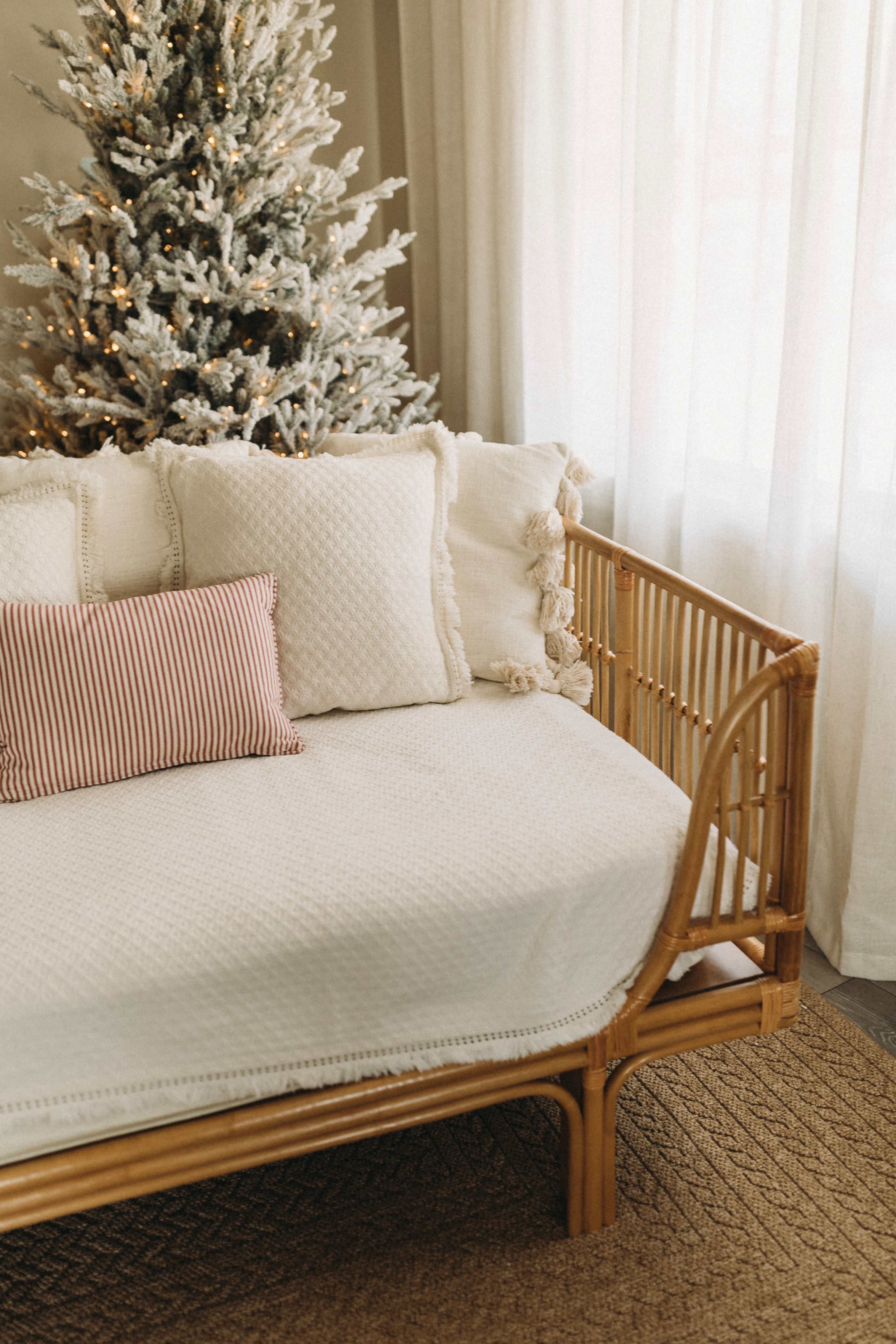 A rattan daybed adorned with a white blanket and decorative pillows sits next to a frosted Christmas tree, with sheer curtains letting in natural light.