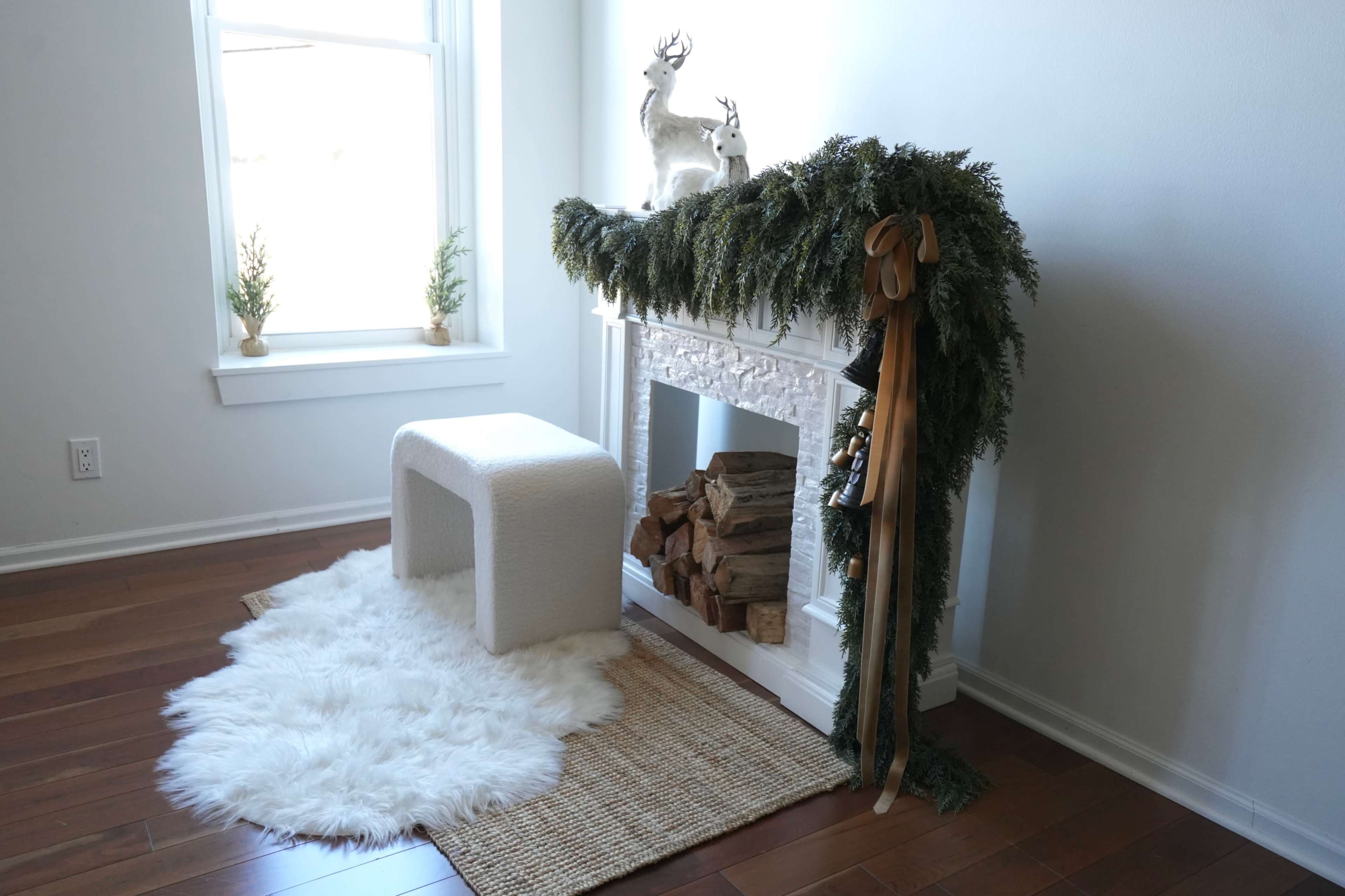 A white faux fur rug is placed in front of a decorative fireplace adorned with greenery and two ornamental deer figurines.