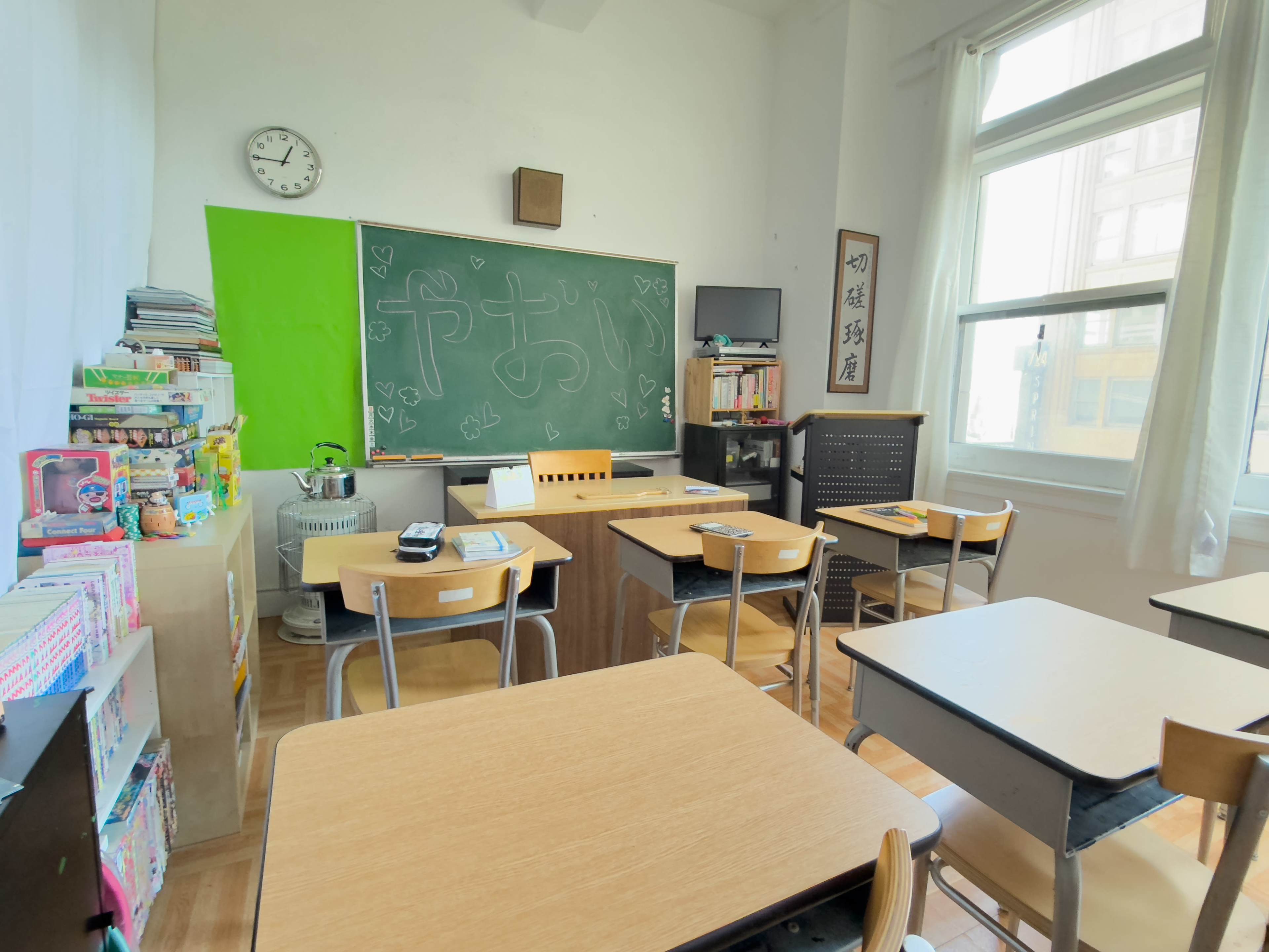 The image shows a classroom with wooden desks arranged around a central blackboard, shelves filled with books and supplies, and a clock on the wall.