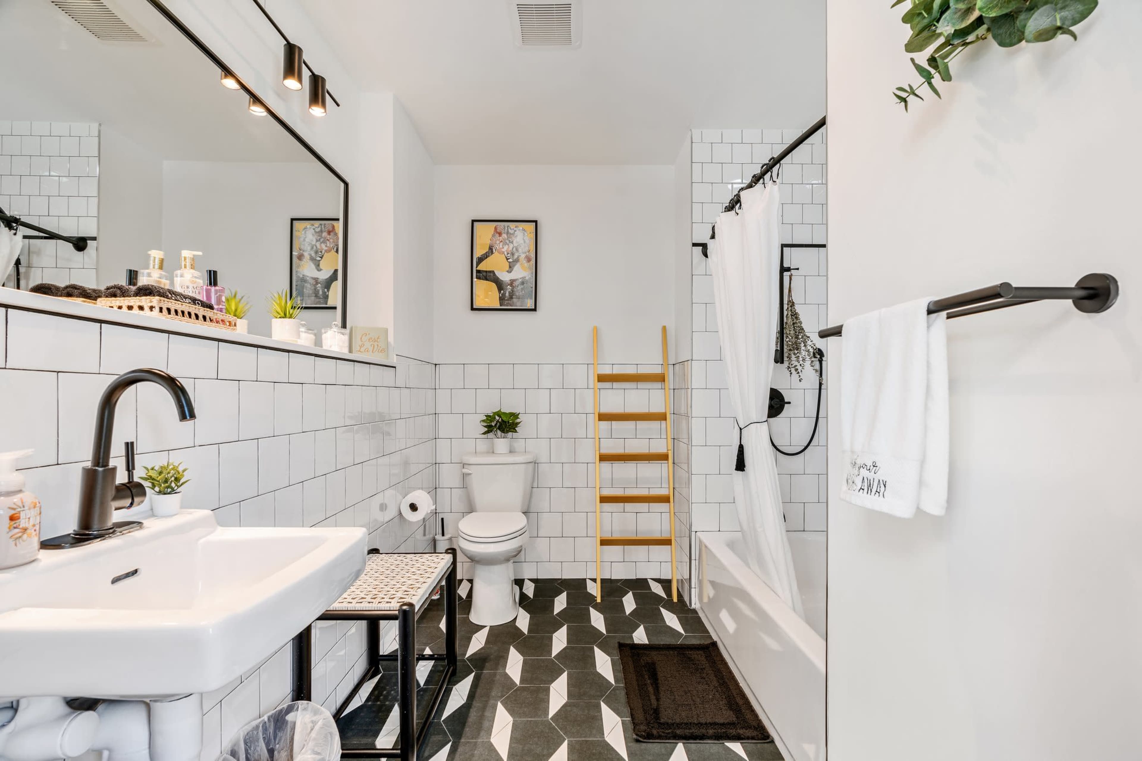The bathroom features white tiled walls, a black and white patterned floor, a bathtub with a shower curtain, a sink with a mirror above it, and decorative plants.