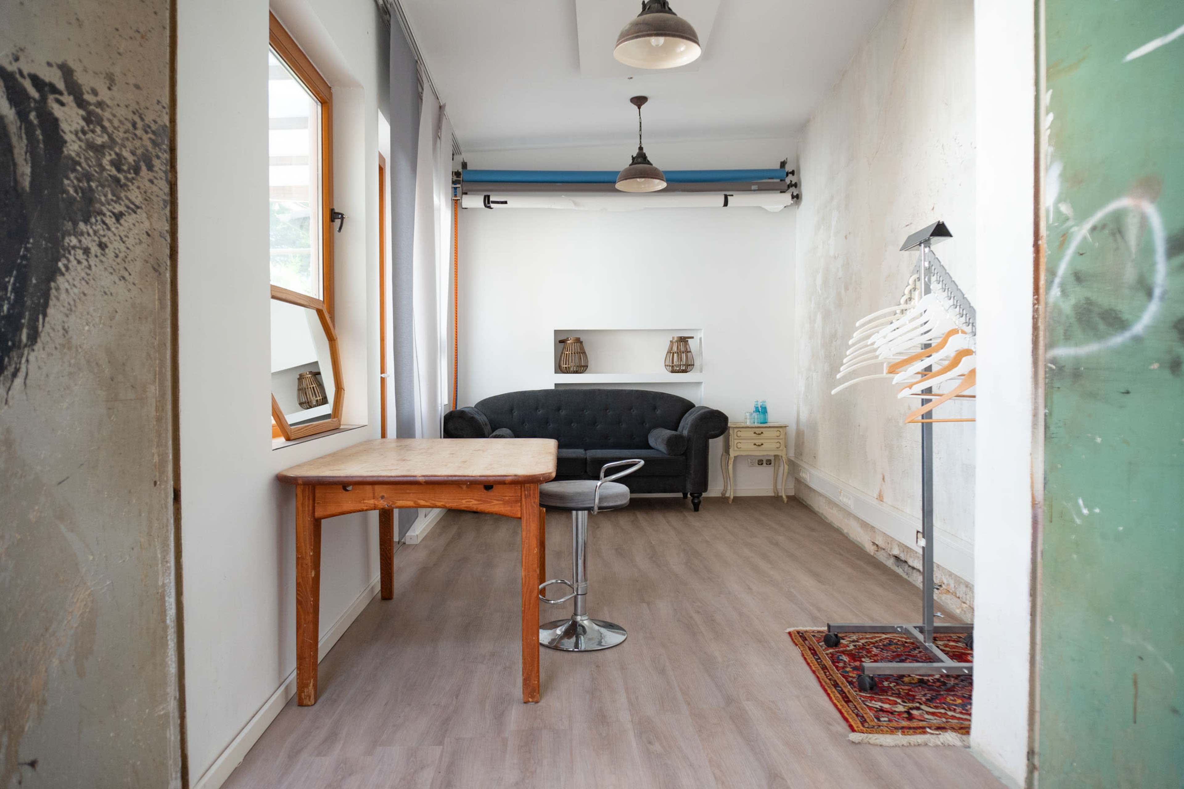 The image shows a minimalist, empty room featuring a wooden table, a black couch, and a clothing rack with hangers.