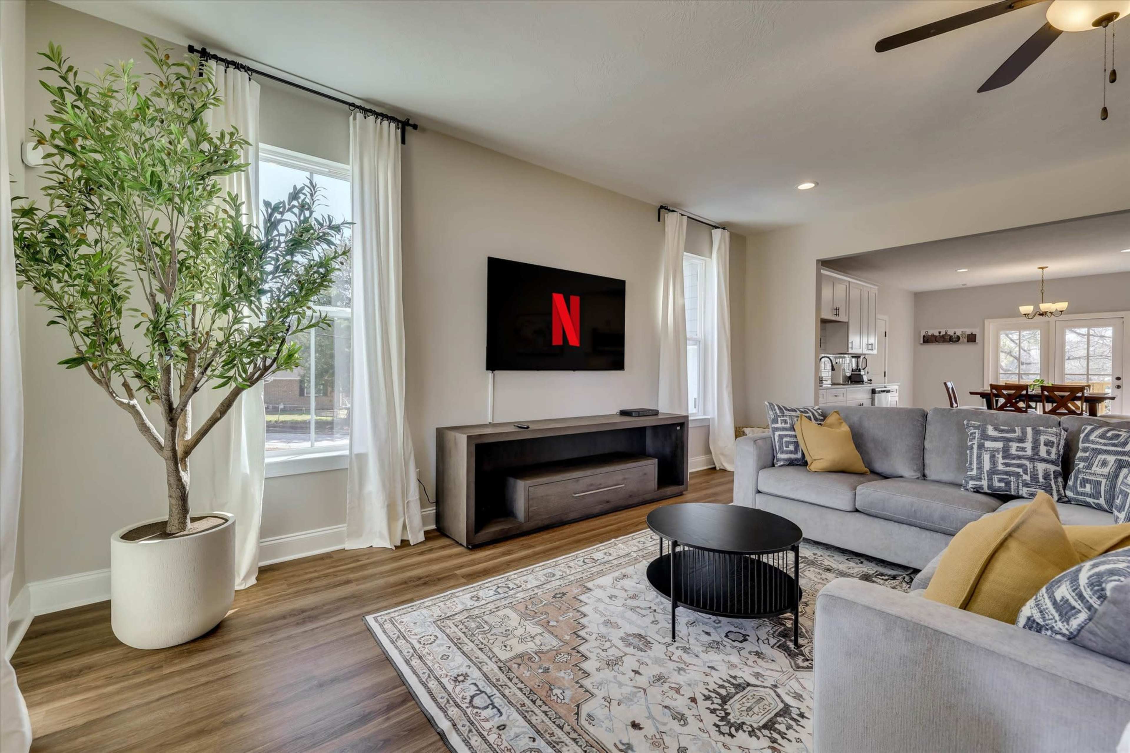 The image shows a modern living room with a large TV mounted on the wall, a gray sectional sofa, a round black coffee table, and a decorative plant by the window, all set against a backdrop of light-colored walls and wood flooring.