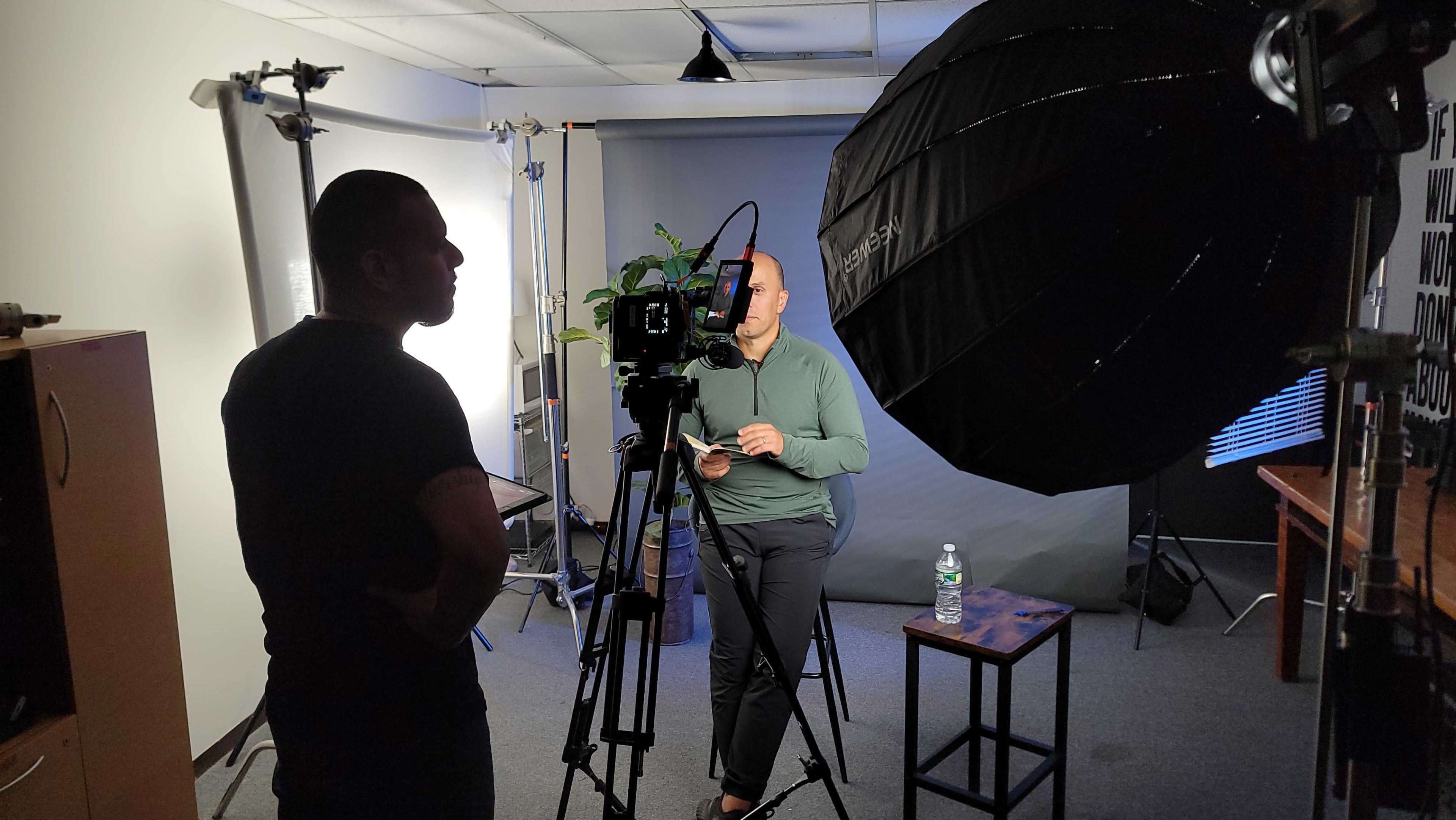 A person sits on a stool in front of a camera and lighting setup, while another individual stands nearby, holding a script.