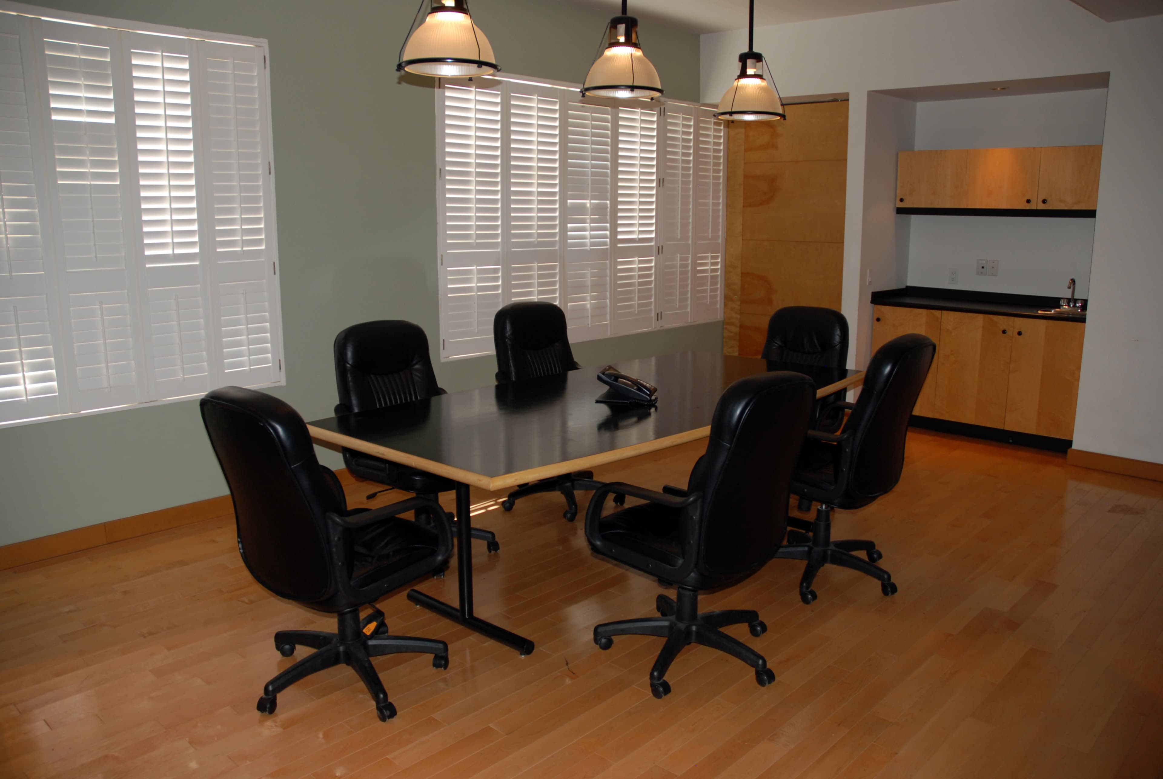 A conference room with a large table surrounded by six black office chairs, natural light filtering in through window blinds, and a kitchenette area in the background.