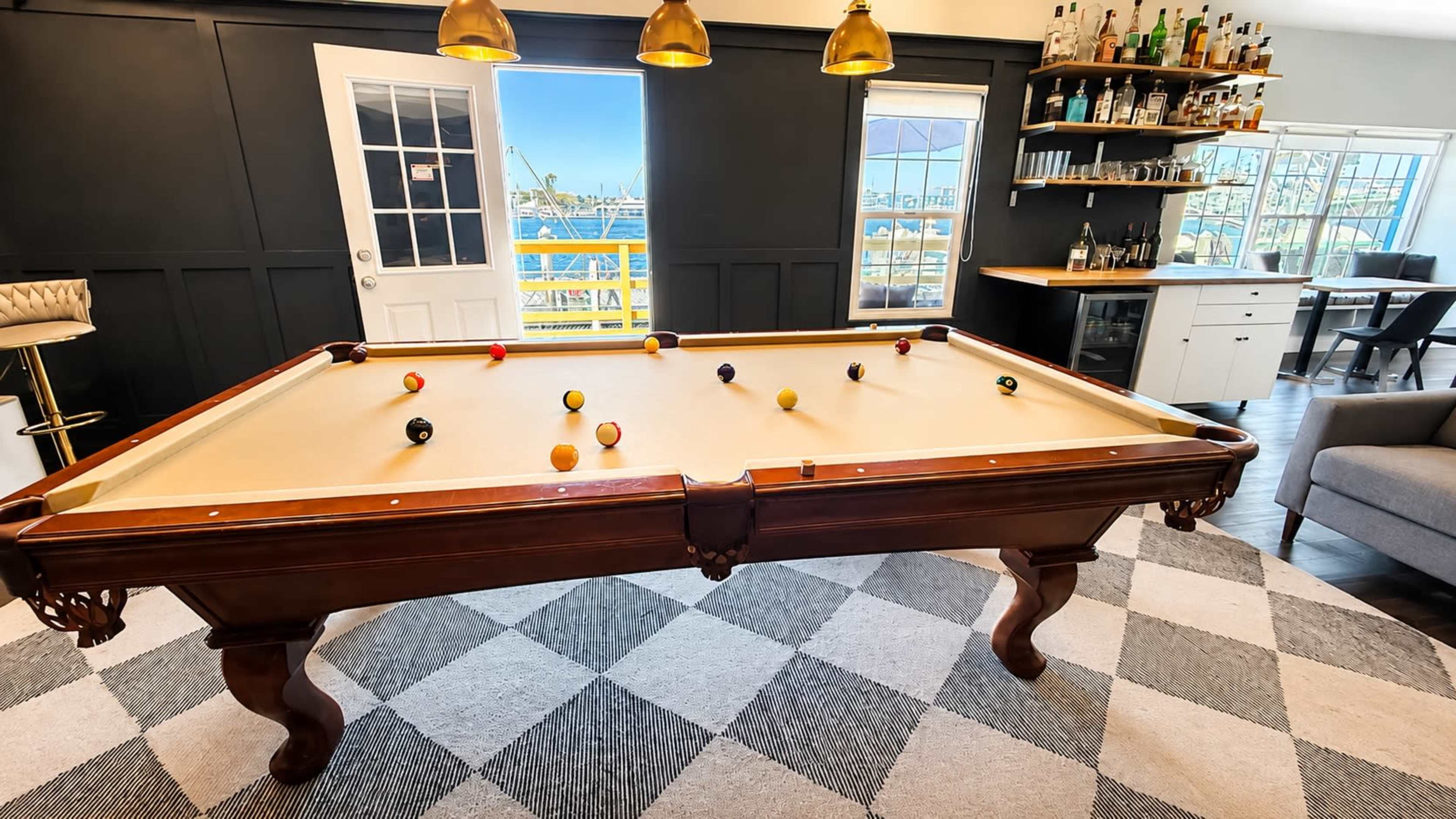 A billiard table with several colored balls is positioned in a room featuring large windows, a bar area, and modern furnishings.