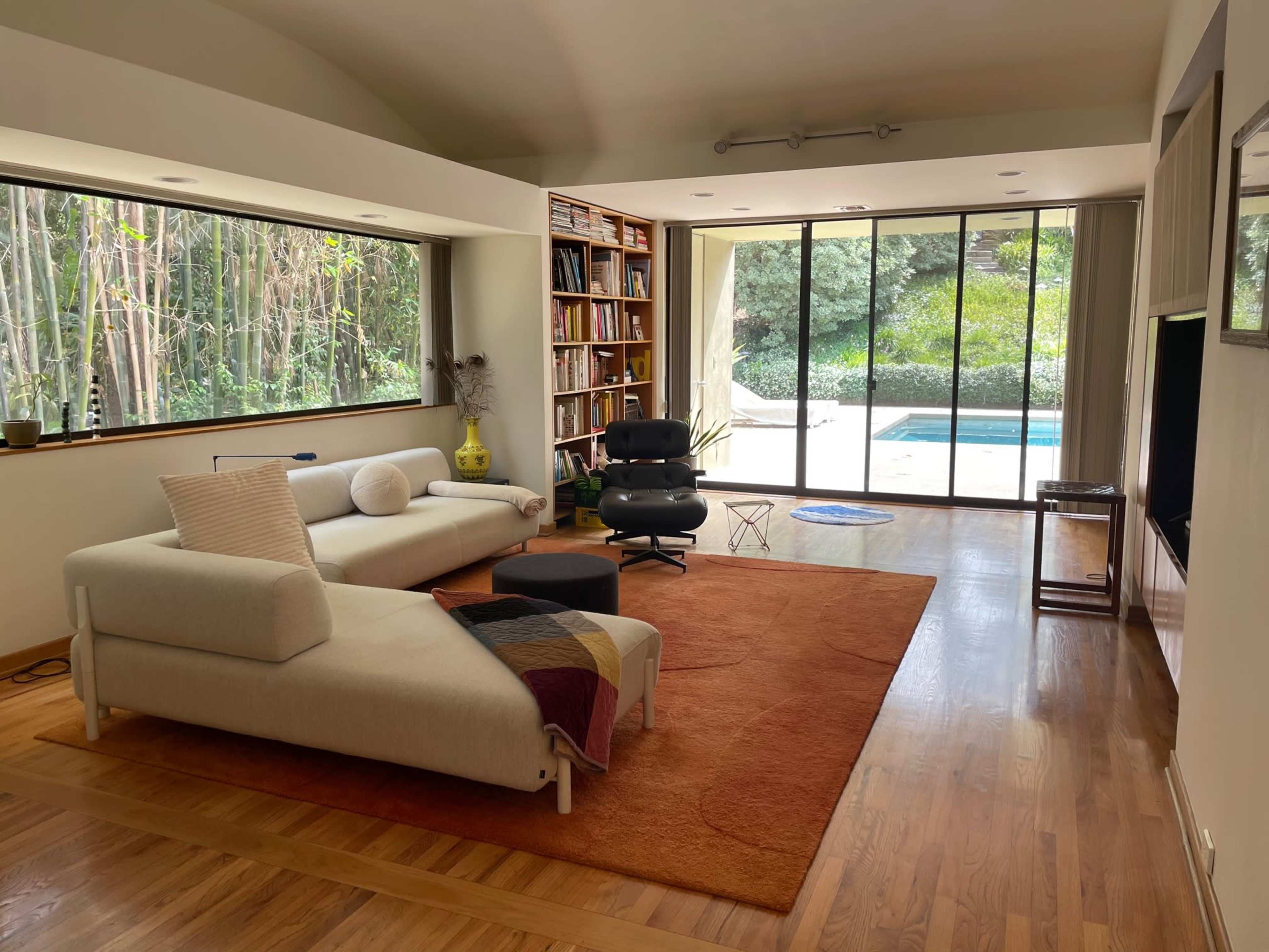 A modern living room features a light-colored couch on a large orange rug, with bookshelves lining one wall and large windows overlooking a pool and greenery outside.