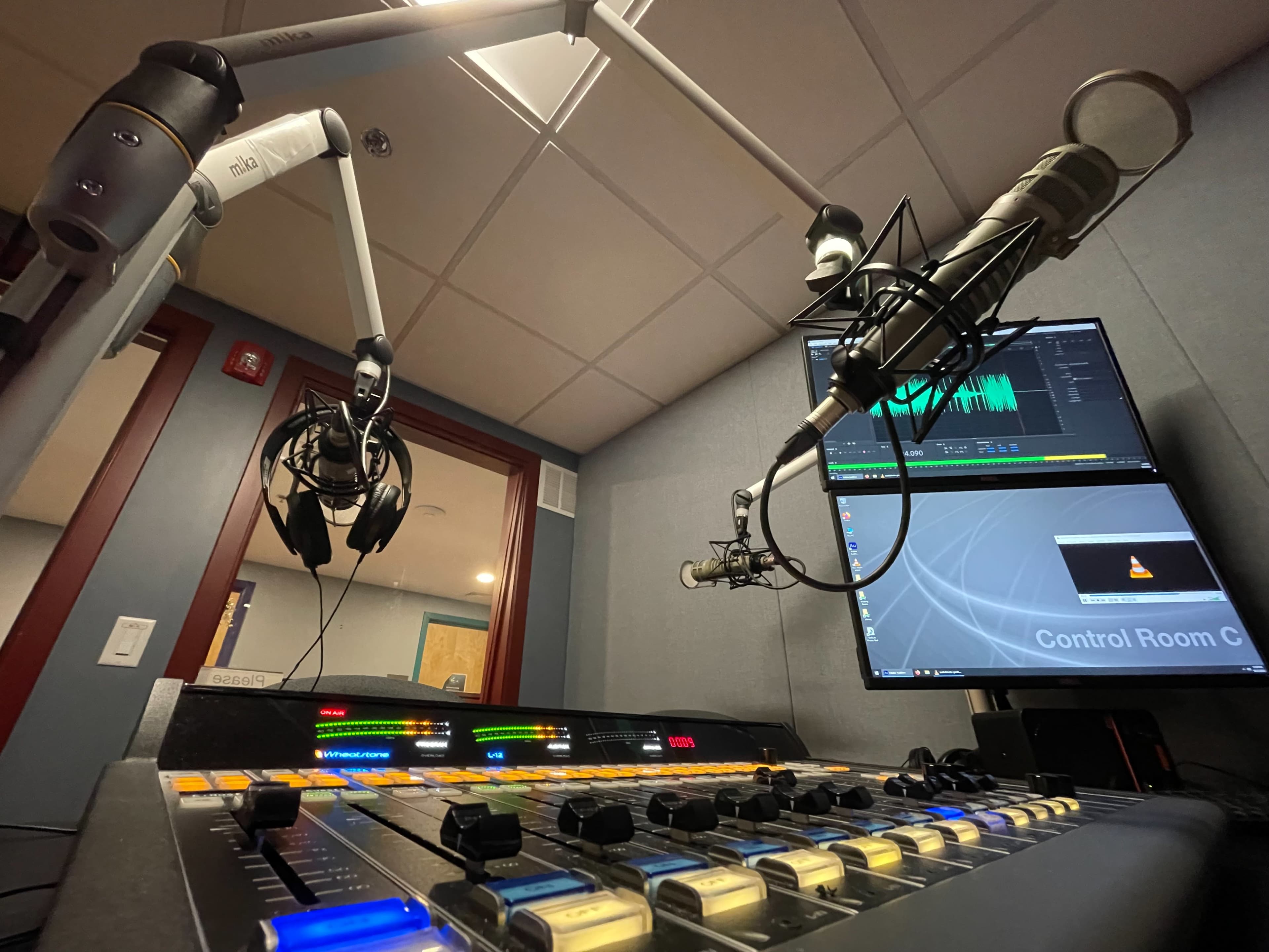 A radio control room with microphones mounted on adjustable arms, a soundboard with various controls, and a computer monitor displaying audio editing software.