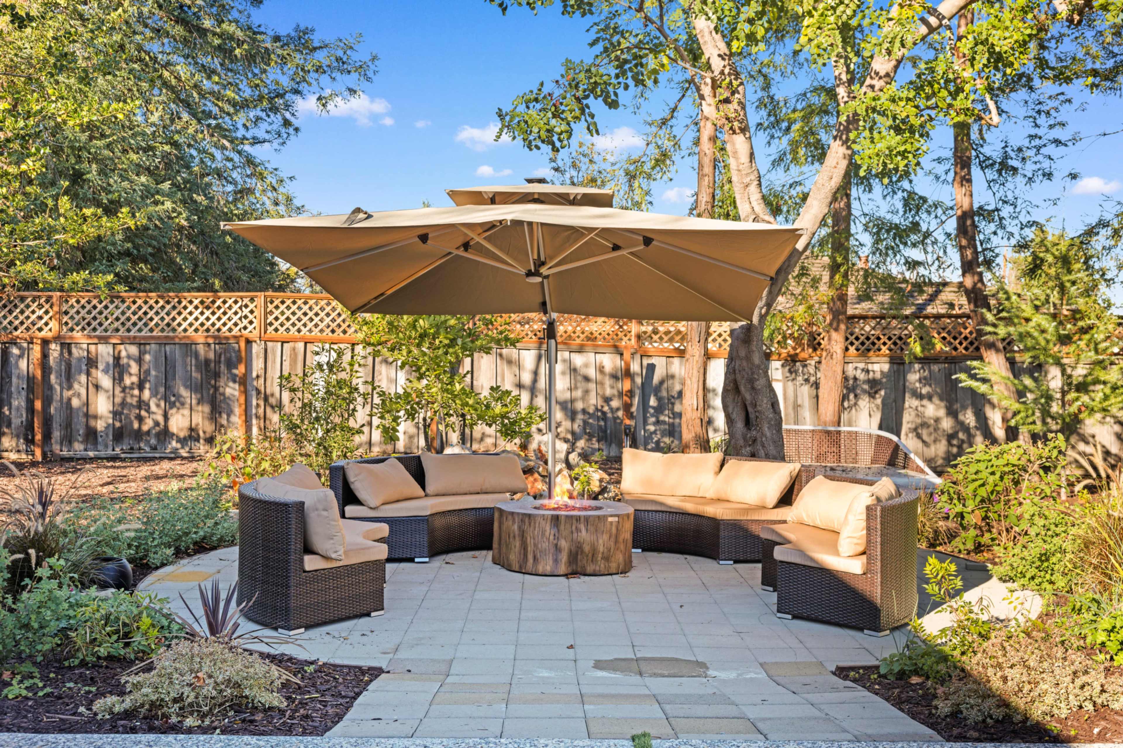 A circular seating area with a tan umbrella and a wooden table is surrounded by plants in a garden.