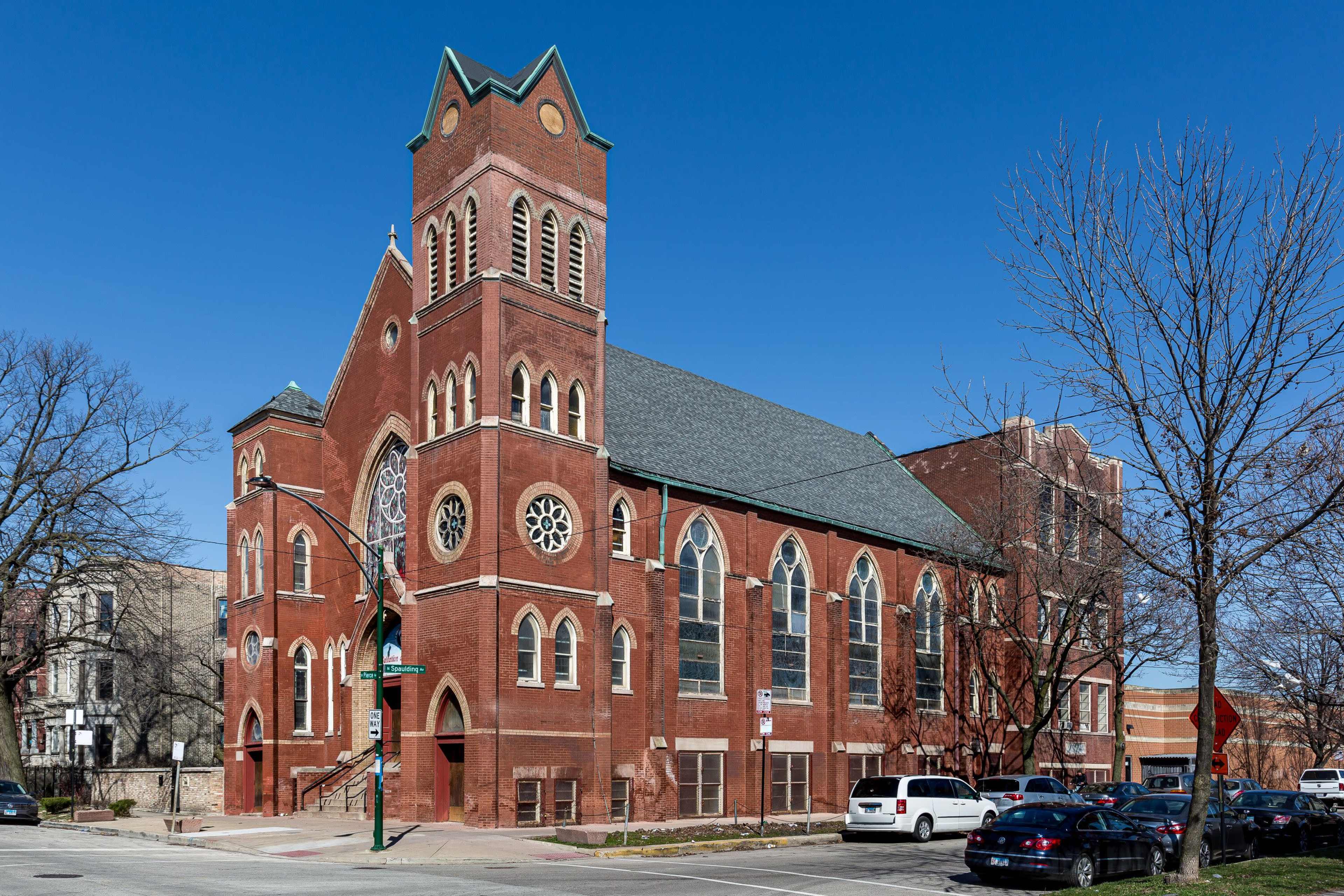 A large red brick church with a tall steeple and distinctive architectural features located at an intersection on a clear blue day.