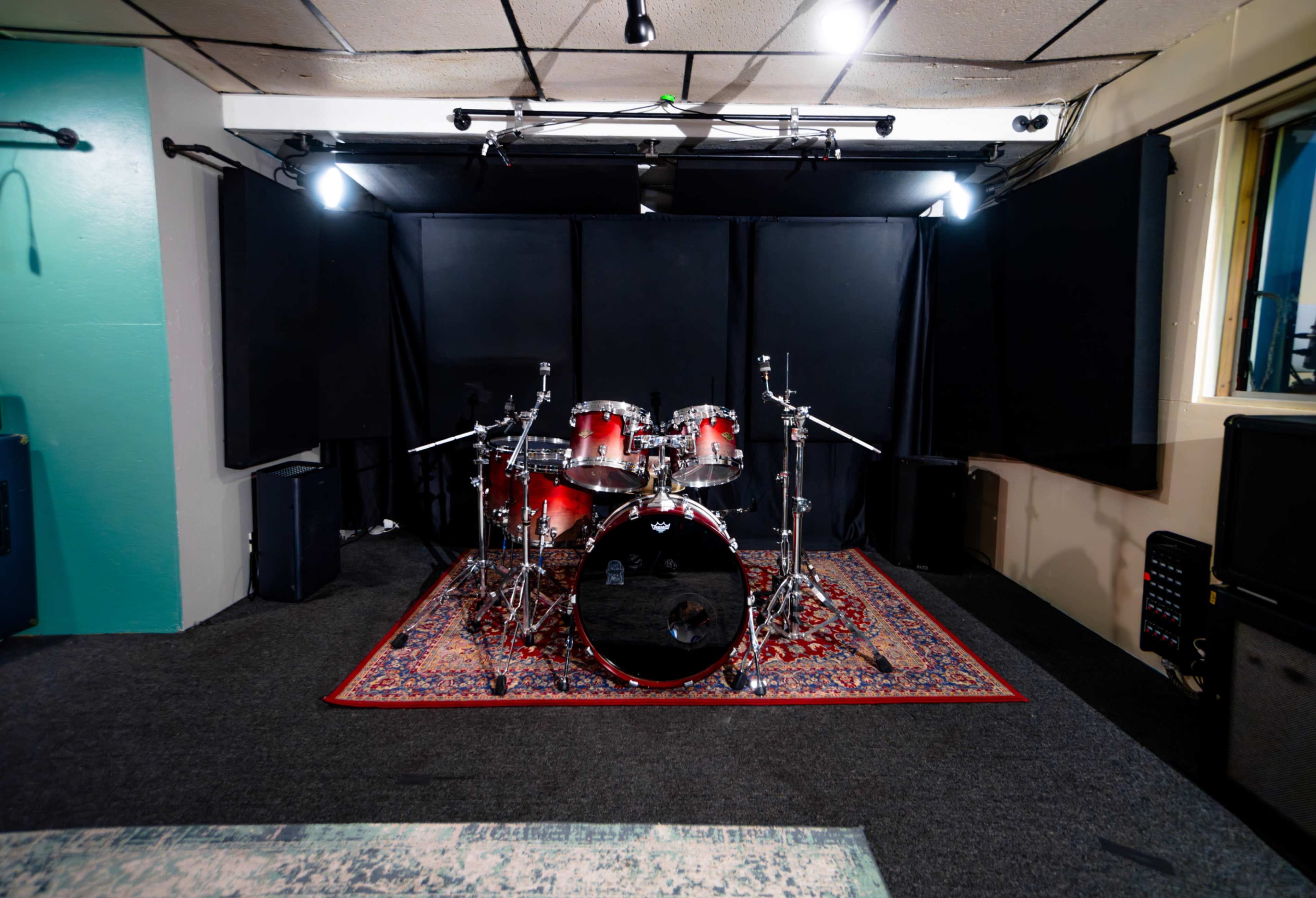 A sound studio with a red drum set positioned on a patterned rug, surrounded by soundproofing panels and equipment on the walls.