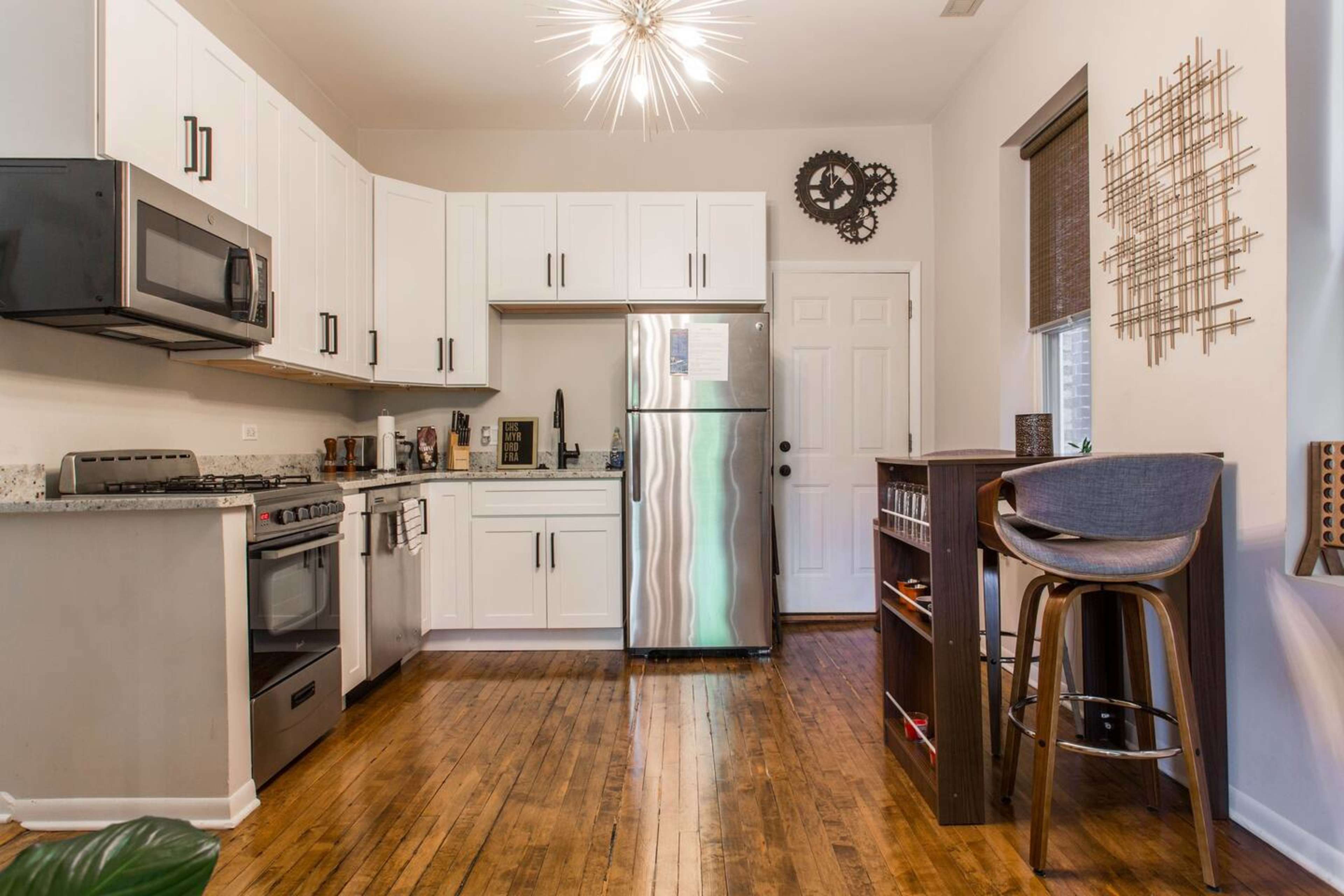 A compact kitchen features white cabinets, stainless steel appliances, and wood flooring, with a bar stool at a small counter.