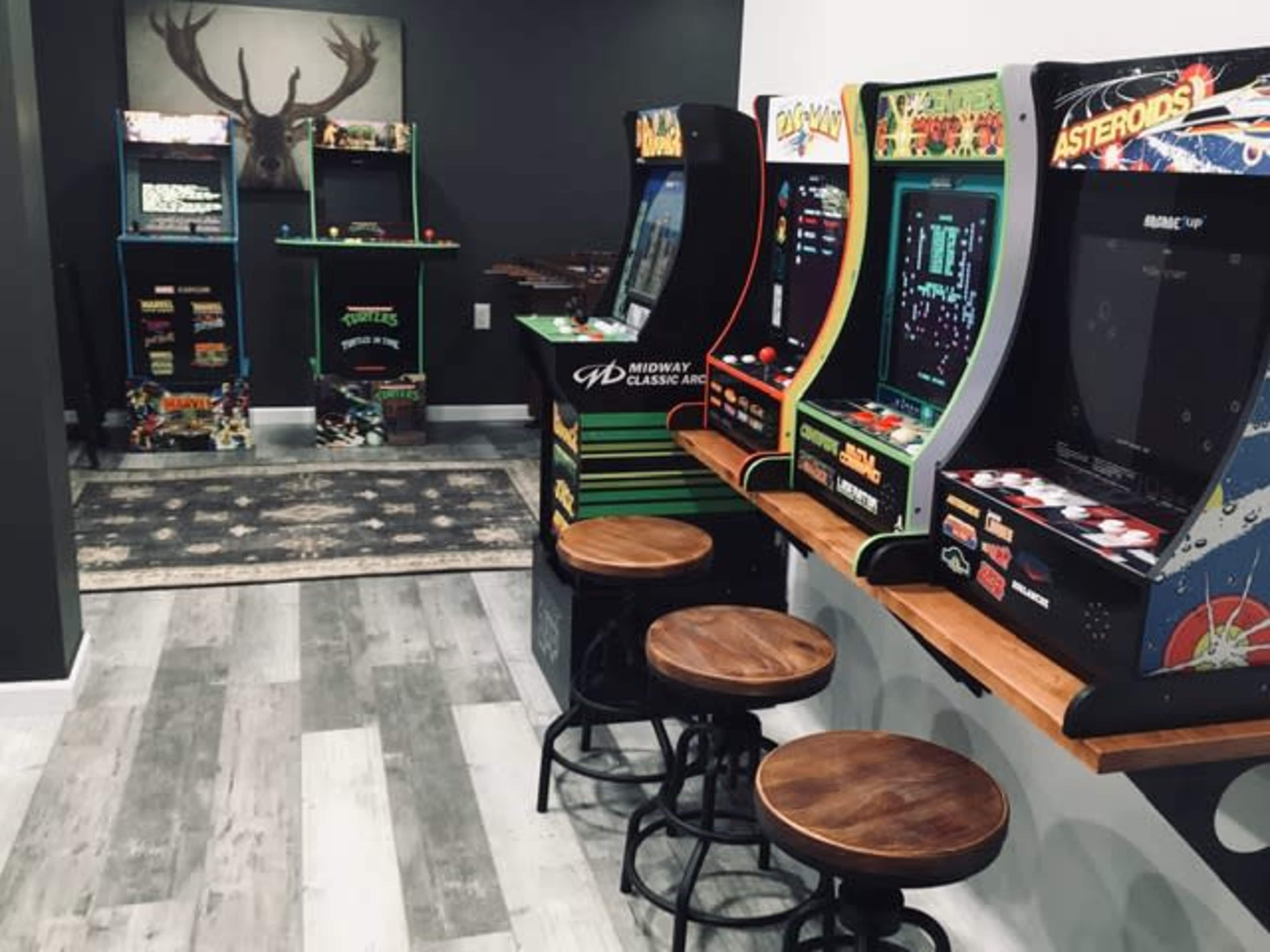 A game room featuring multiple retro arcade machines lined up along one wall, accompanied by wooden stools for seating.