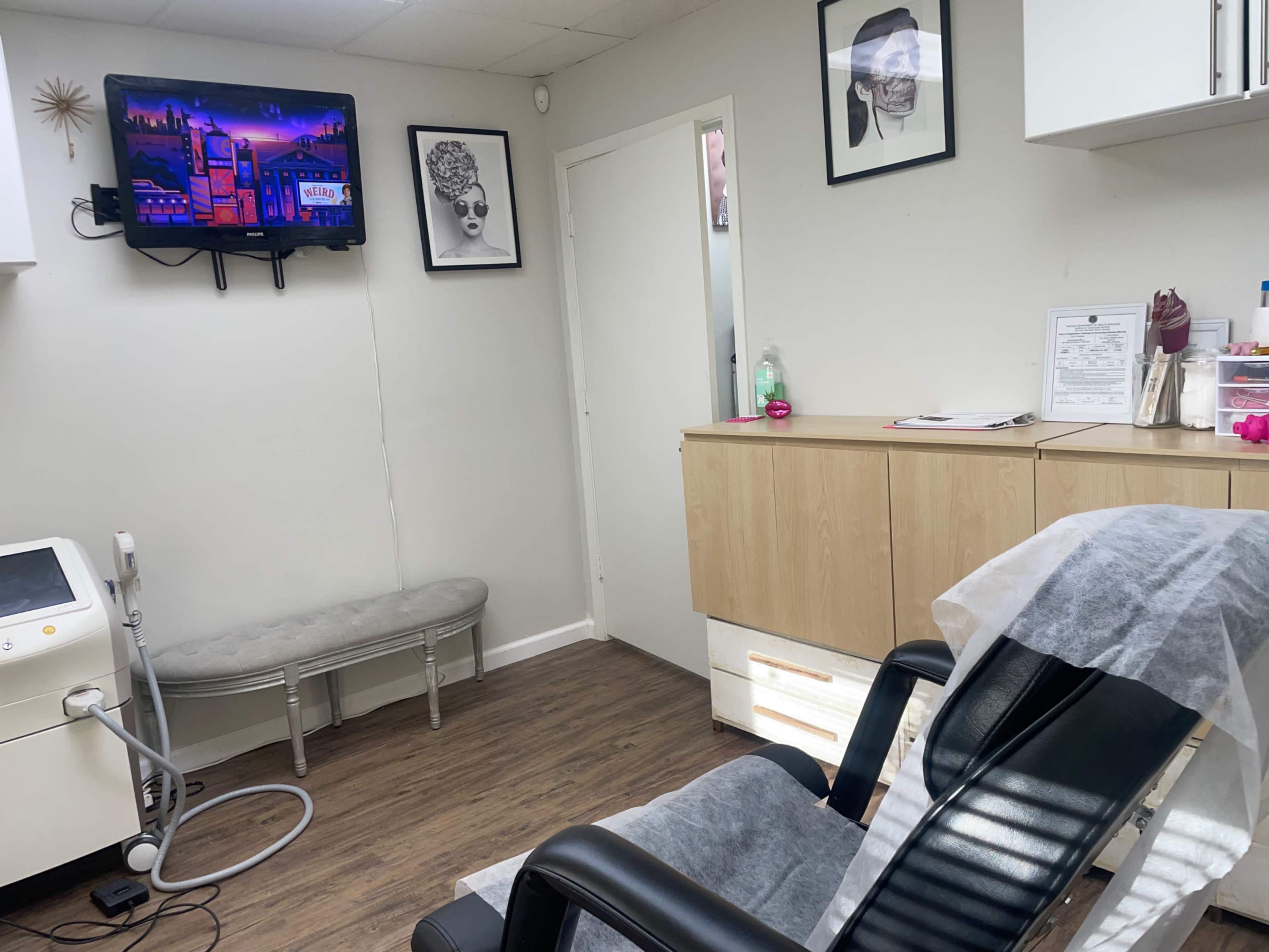 A small treatment room with a black reclining chair, a wooden cabinet, a television mounted on the wall, and a decorative wall featuring two framed pictures.