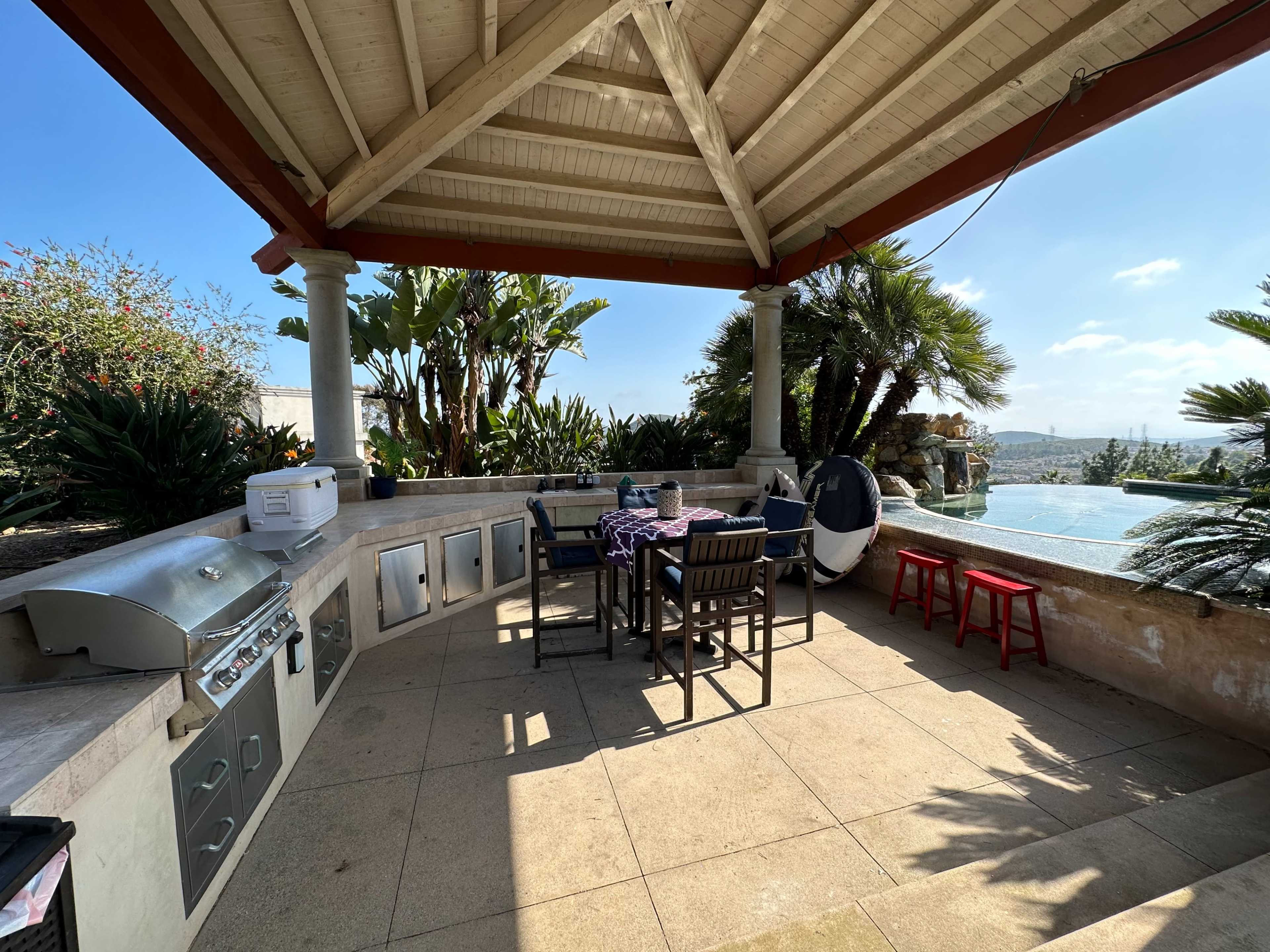 The image shows an outdoor patio area with a barbecue grill, a dining table with chairs, and a view of a swimming pool surrounded by tropical plants.