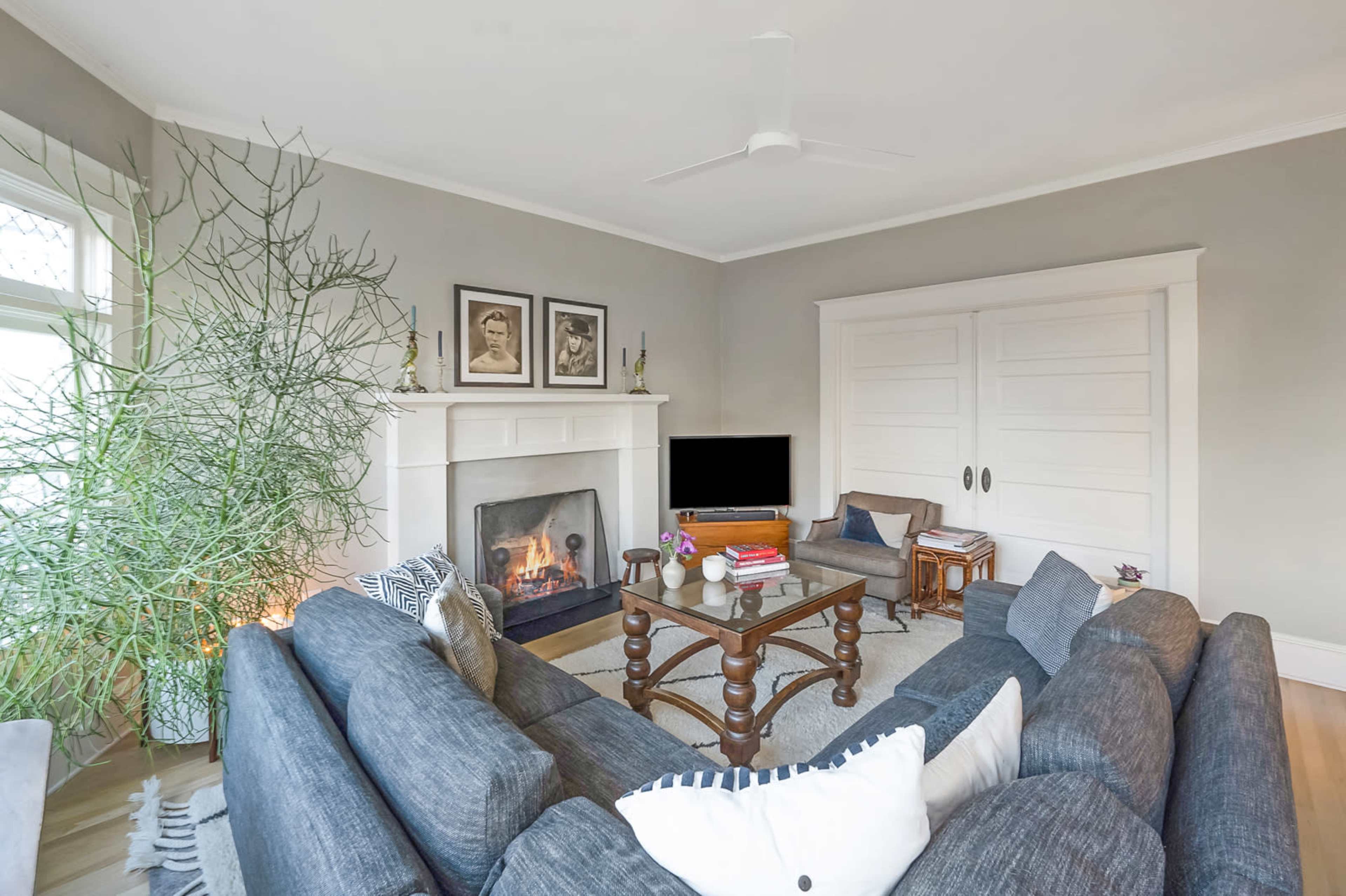 A living room features a gray sofa, a coffee table, a fireplace, and a television against a light-colored wall with framed pictures.