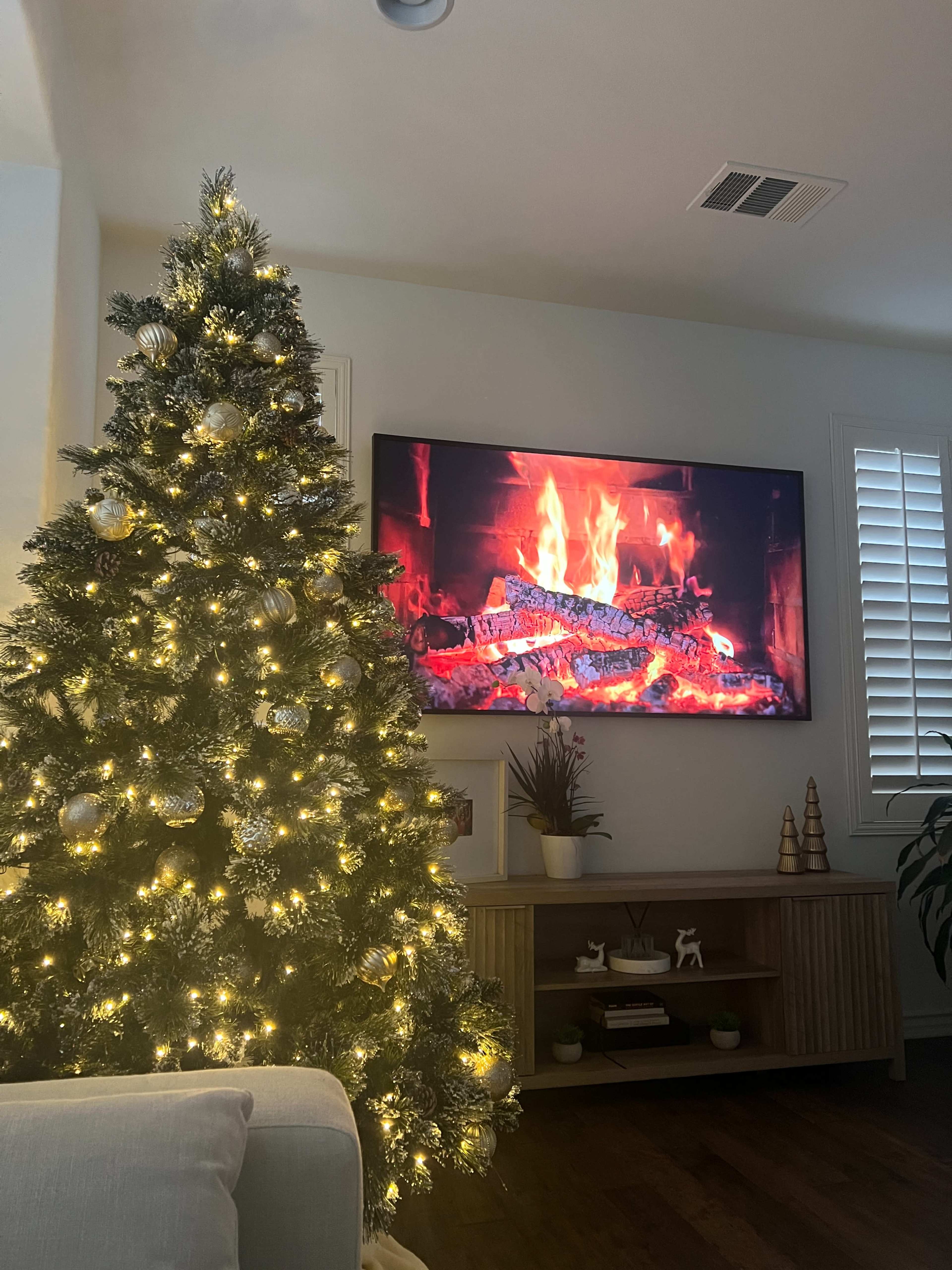 A decorated Christmas tree with lights stands next to a television displaying a fireplace scene in a living room.