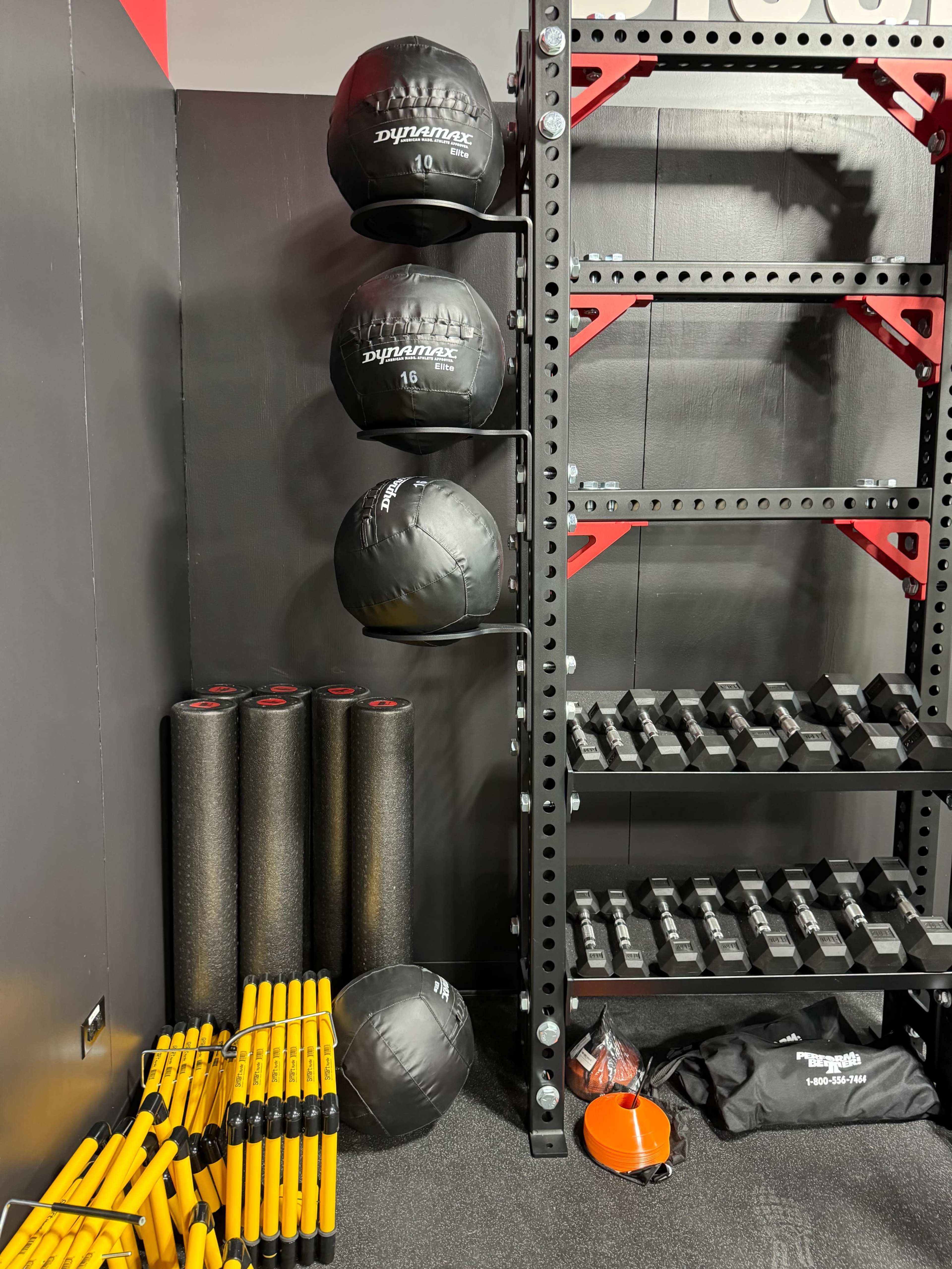 The image shows a gym corner featuring a tall rack holding four black medicine balls and several weights, with foam rollers and training sticks neatly arranged on the floor.