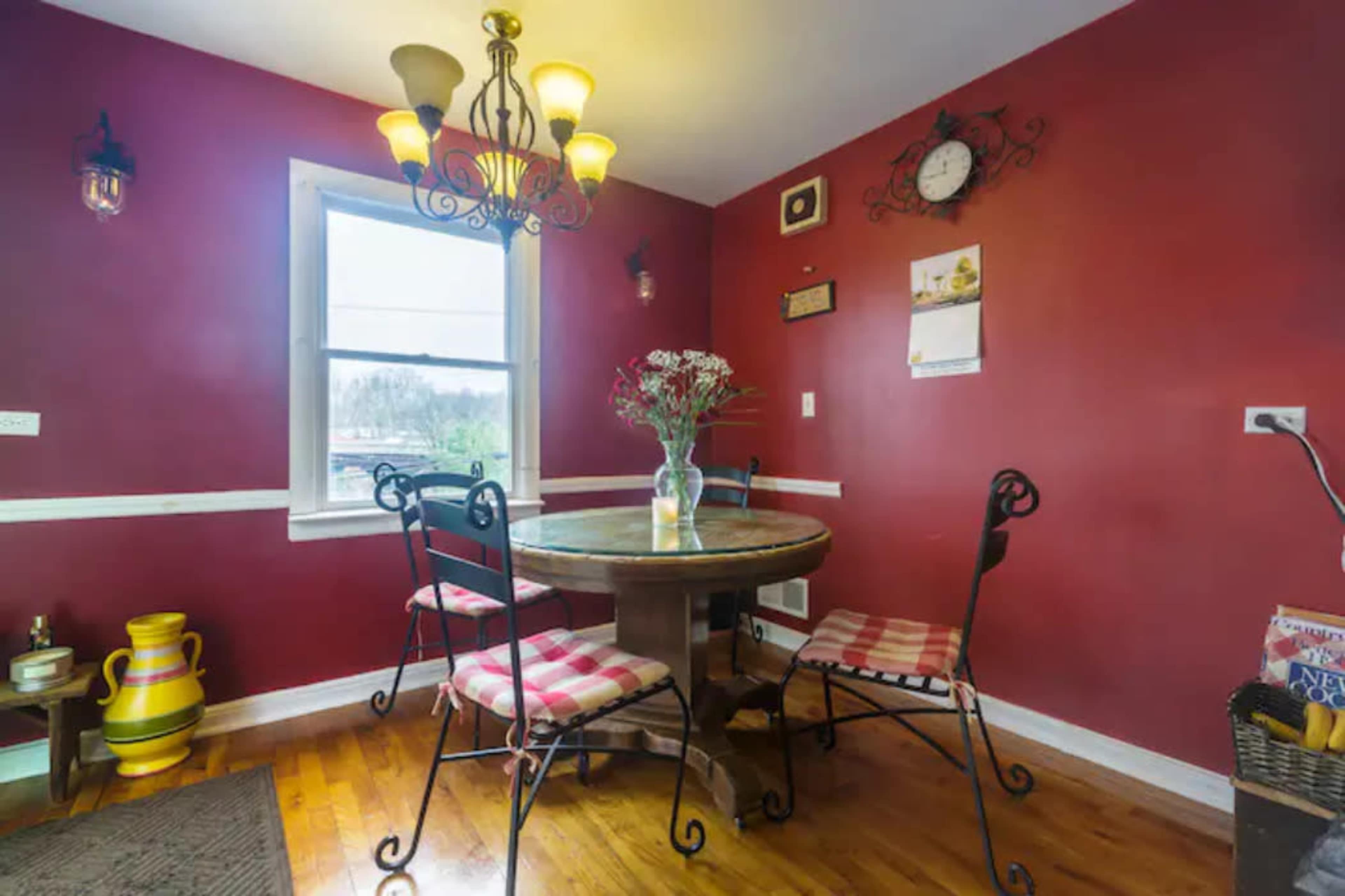 A round wooden dining table surrounded by four metal chairs is set up under a chandelier in a room with red walls.