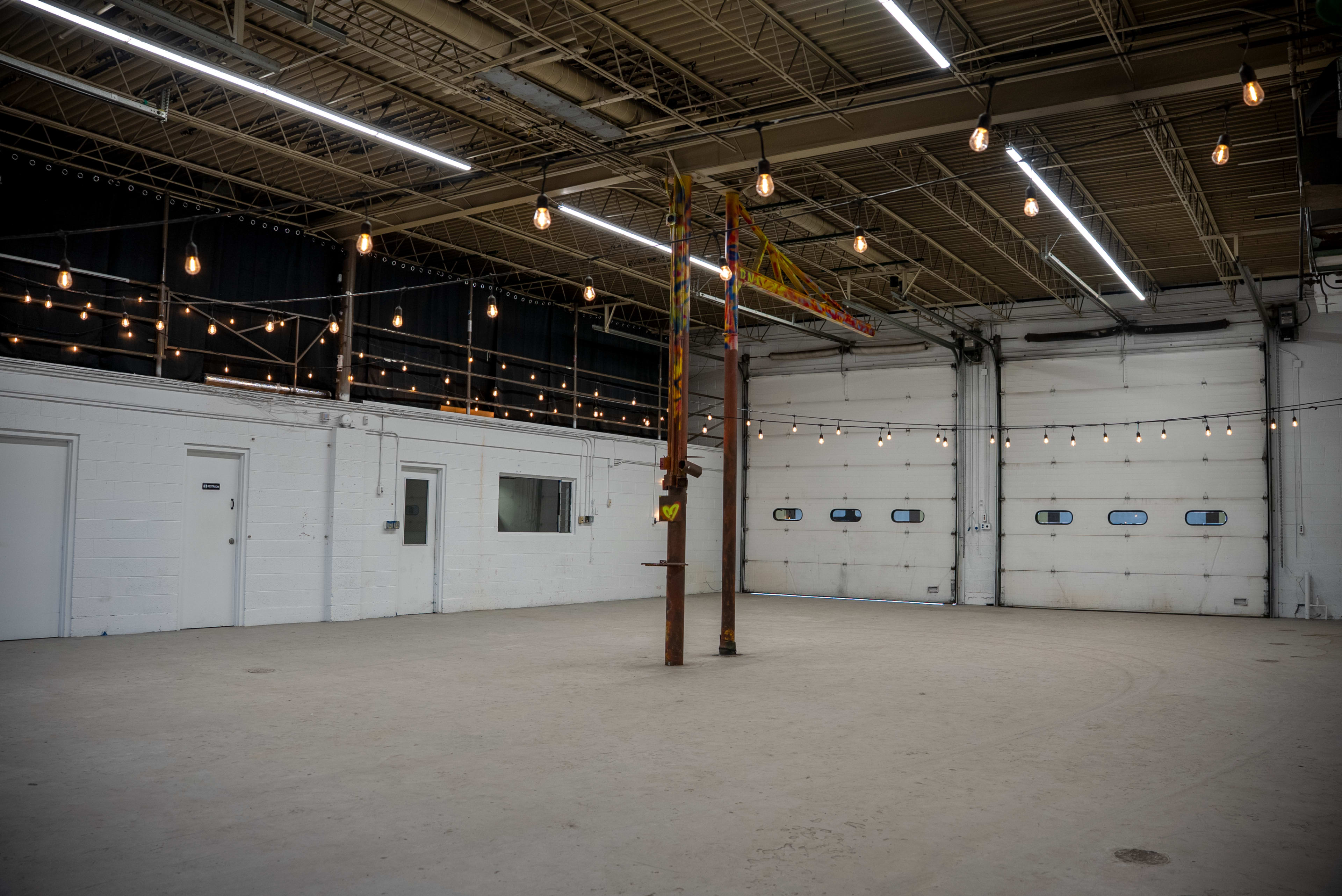 The image shows a large, empty industrial space with white walls, a concrete floor, and overhead lights hanging from the ceiling.