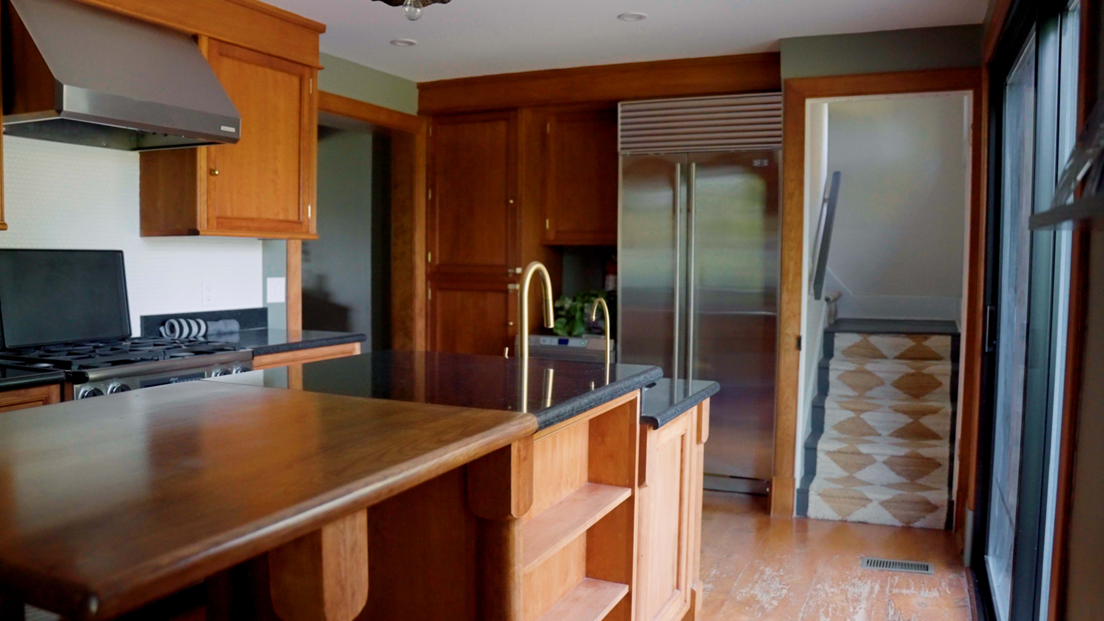 The image shows a kitchen with wooden cabinetry, a central island with open shelving, and a stainless steel refrigerator in the background.