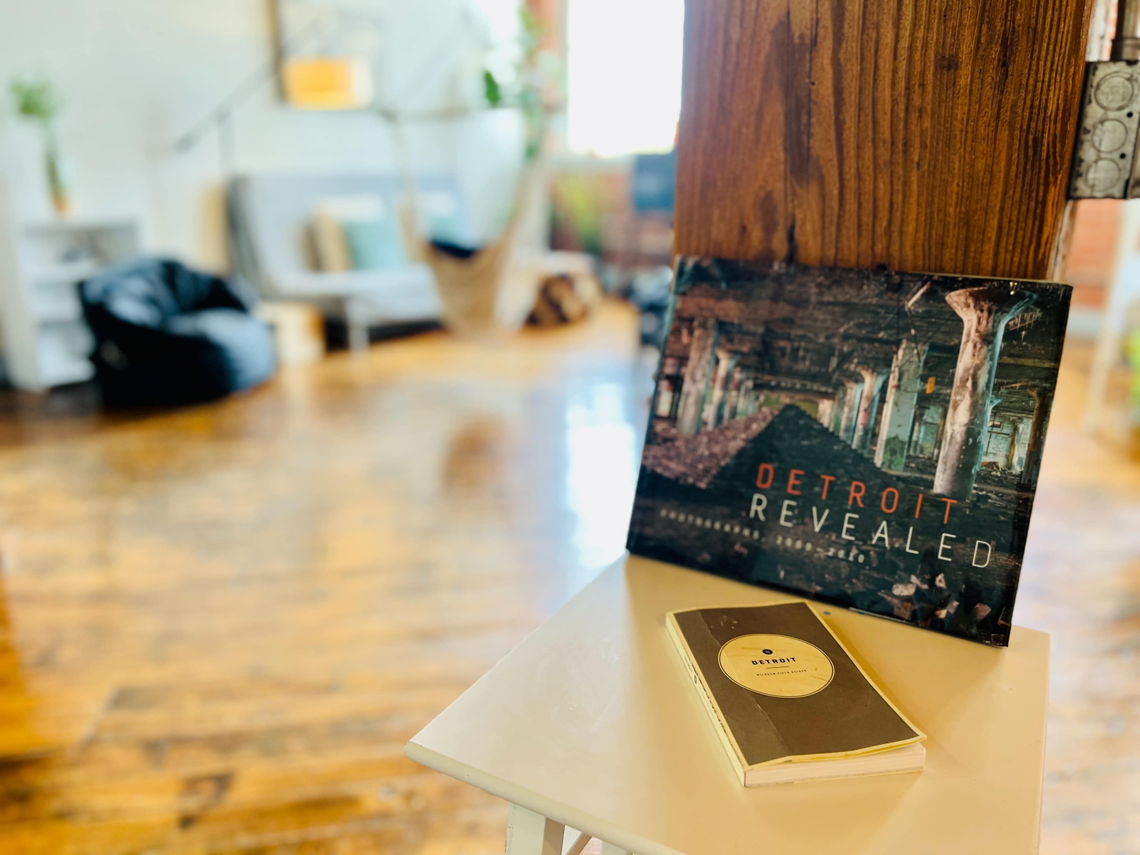 A coffee table holds a magazine titled "Detroit Revealed" next to a notebook in a room featuring hardwood floors and minimal furniture.