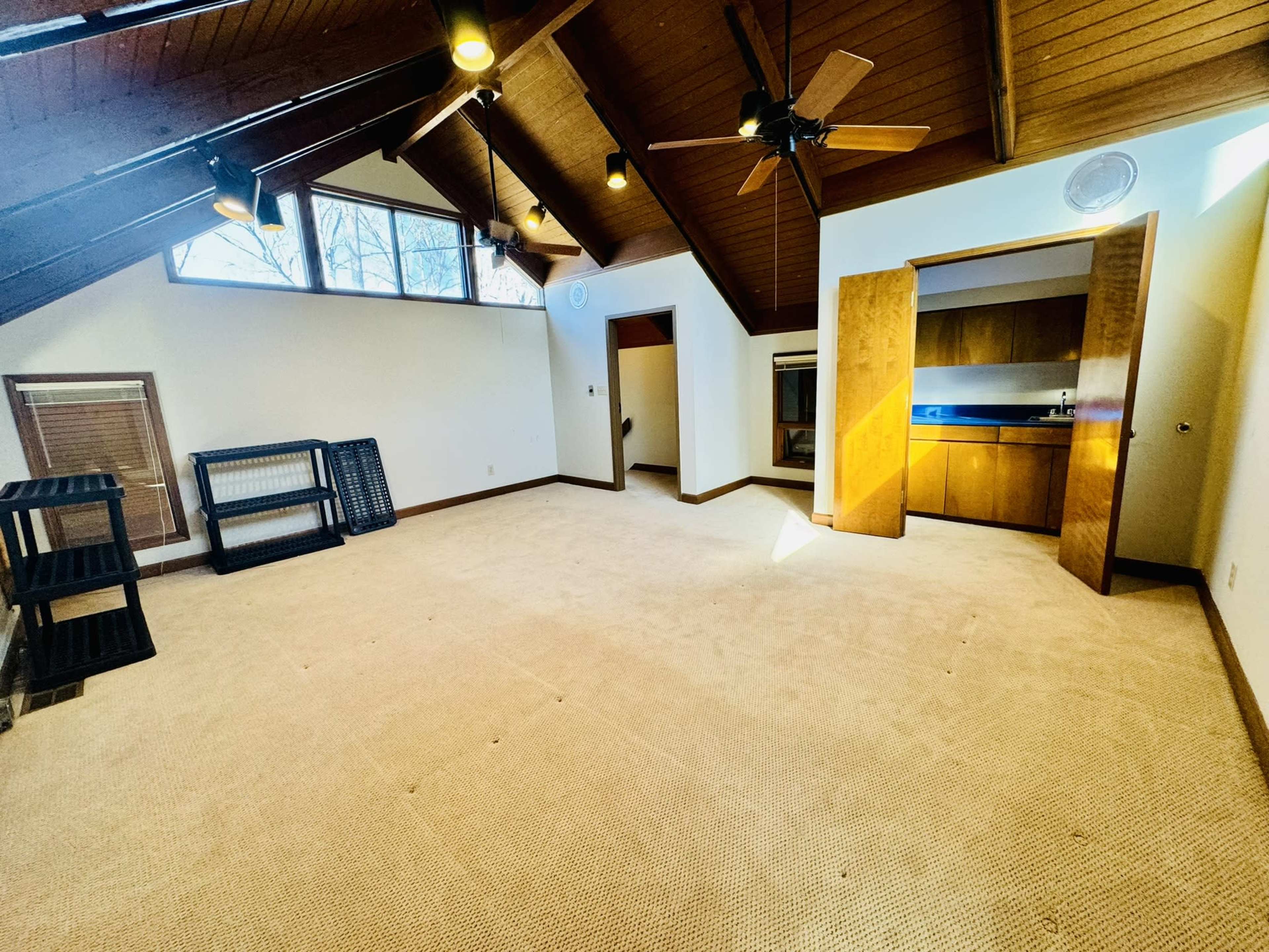 The room features a peaked ceiling with wooden beams, a plush carpeted floor, and a kitchenette area accessible through an opening.
