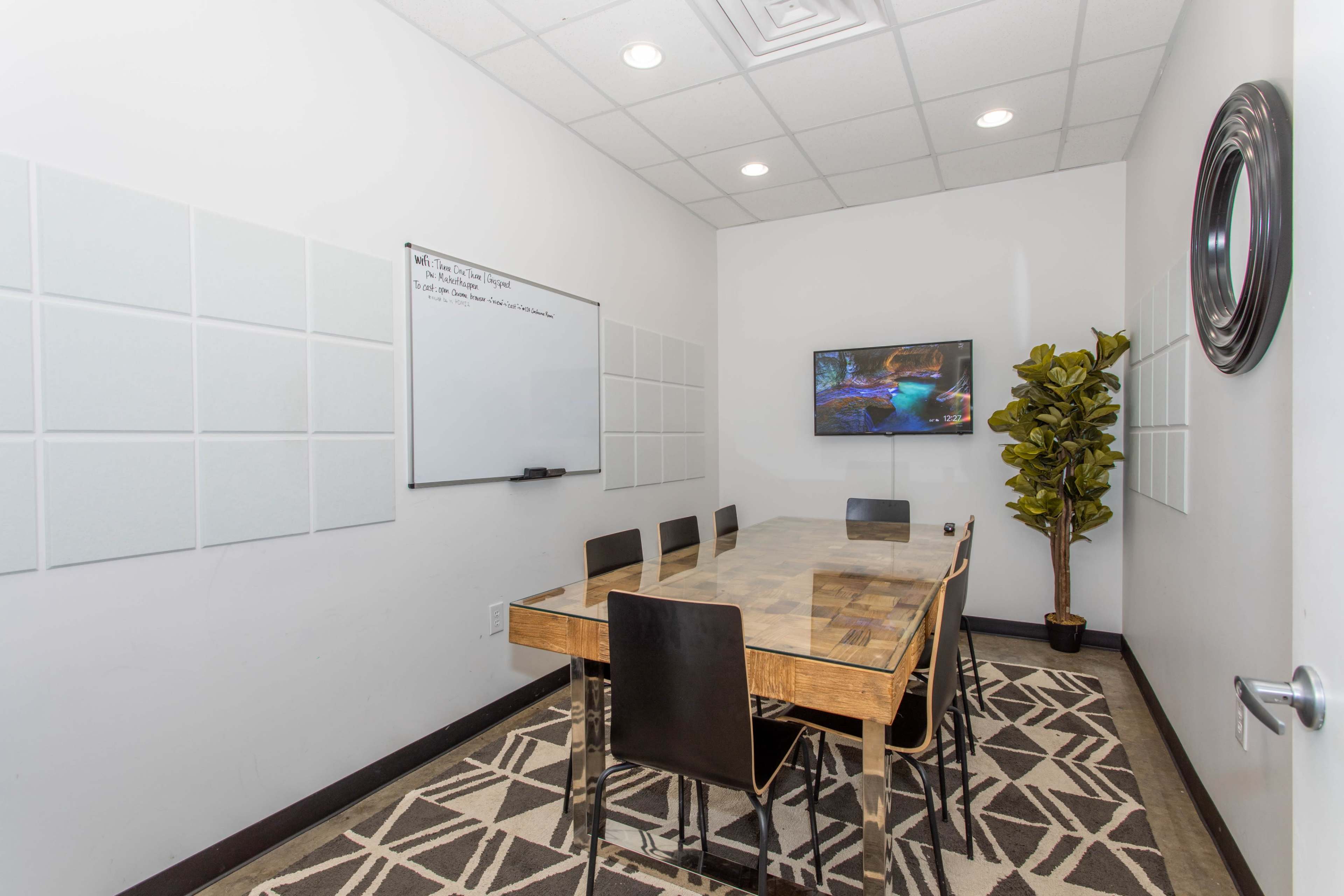 A small conference room features a wooden table surrounded by black chairs, a whiteboard on one wall, and a television mounted on another wall, alongside a decorative plant.