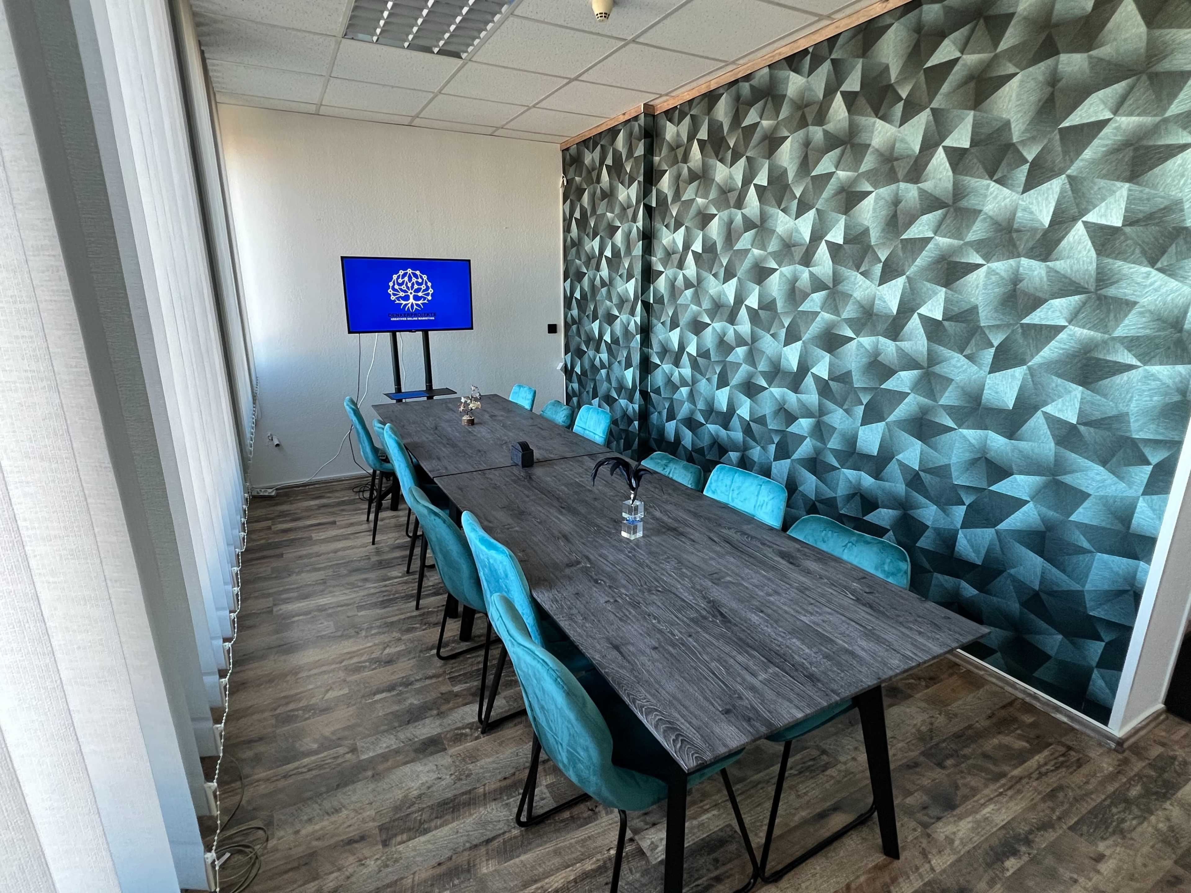 A modern conference room features a long wooden table surrounded by blue chairs, with a large screen displaying a logo on one wall.