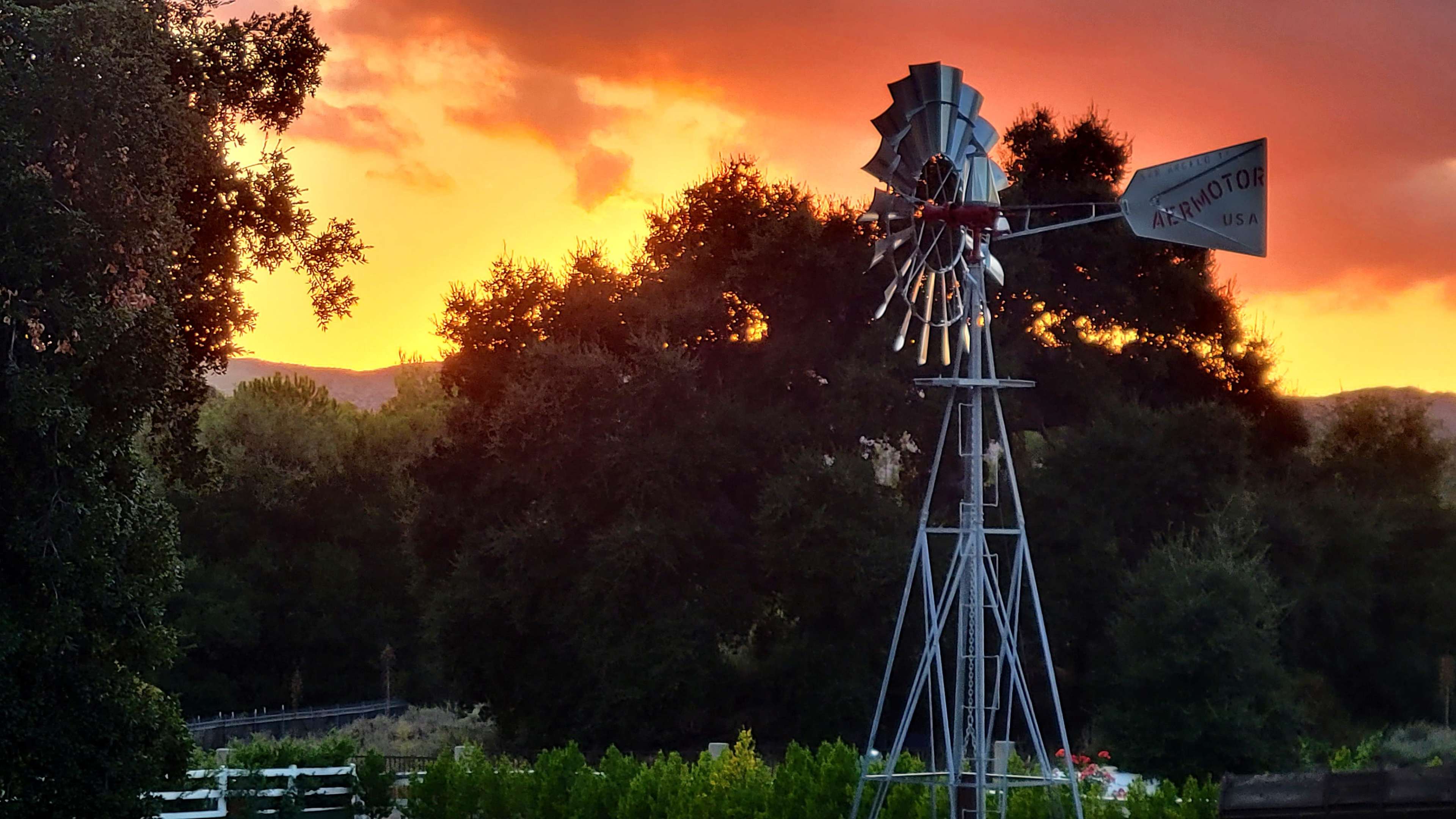 A metal windmill stands against a colorful sunset, with dark trees in the background.