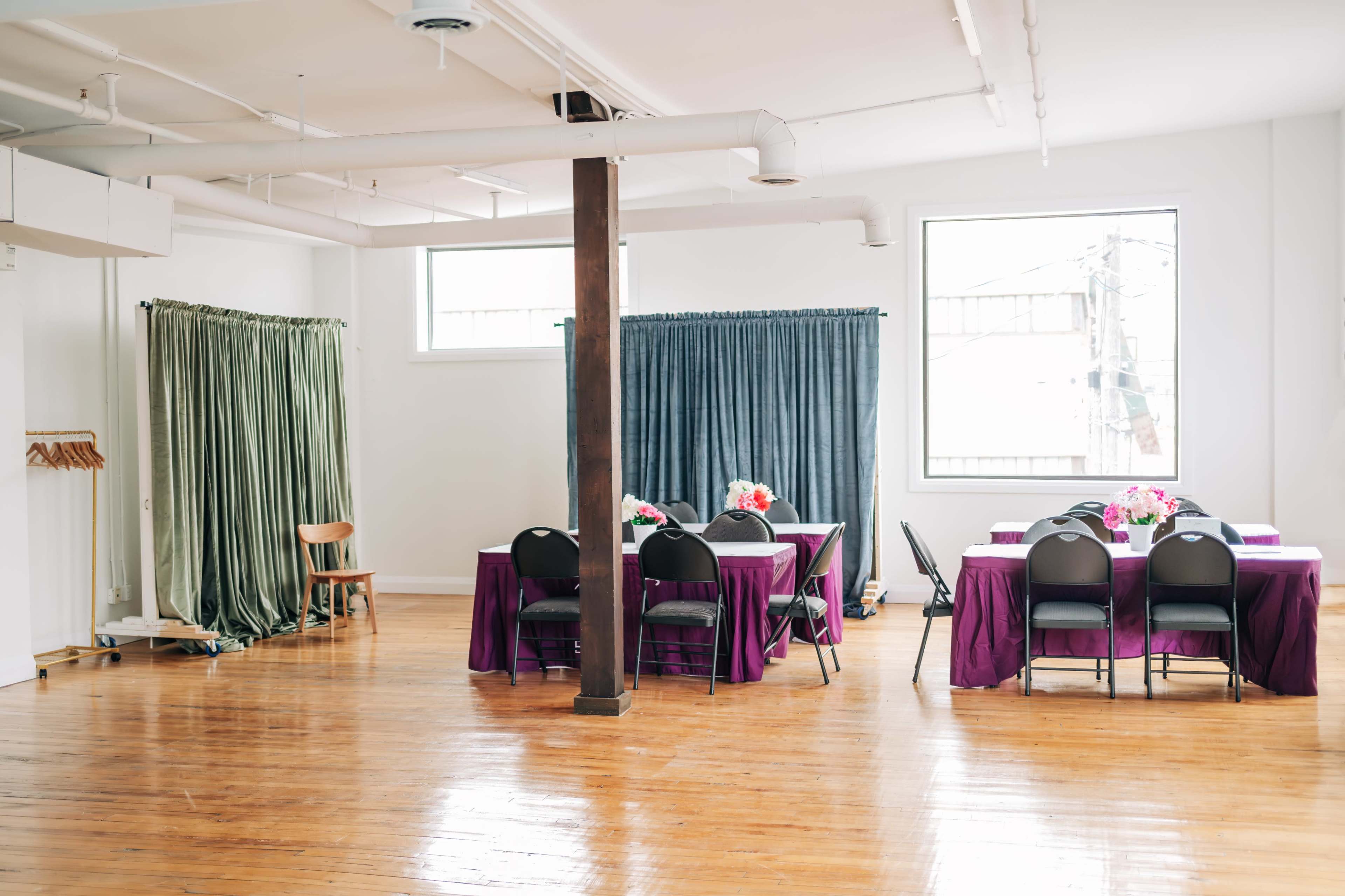 A spacious room with wooden flooring, featuring two clusters of tables covered in colorful tablecloths and surrounded by chairs, along with curtains on the windows.
