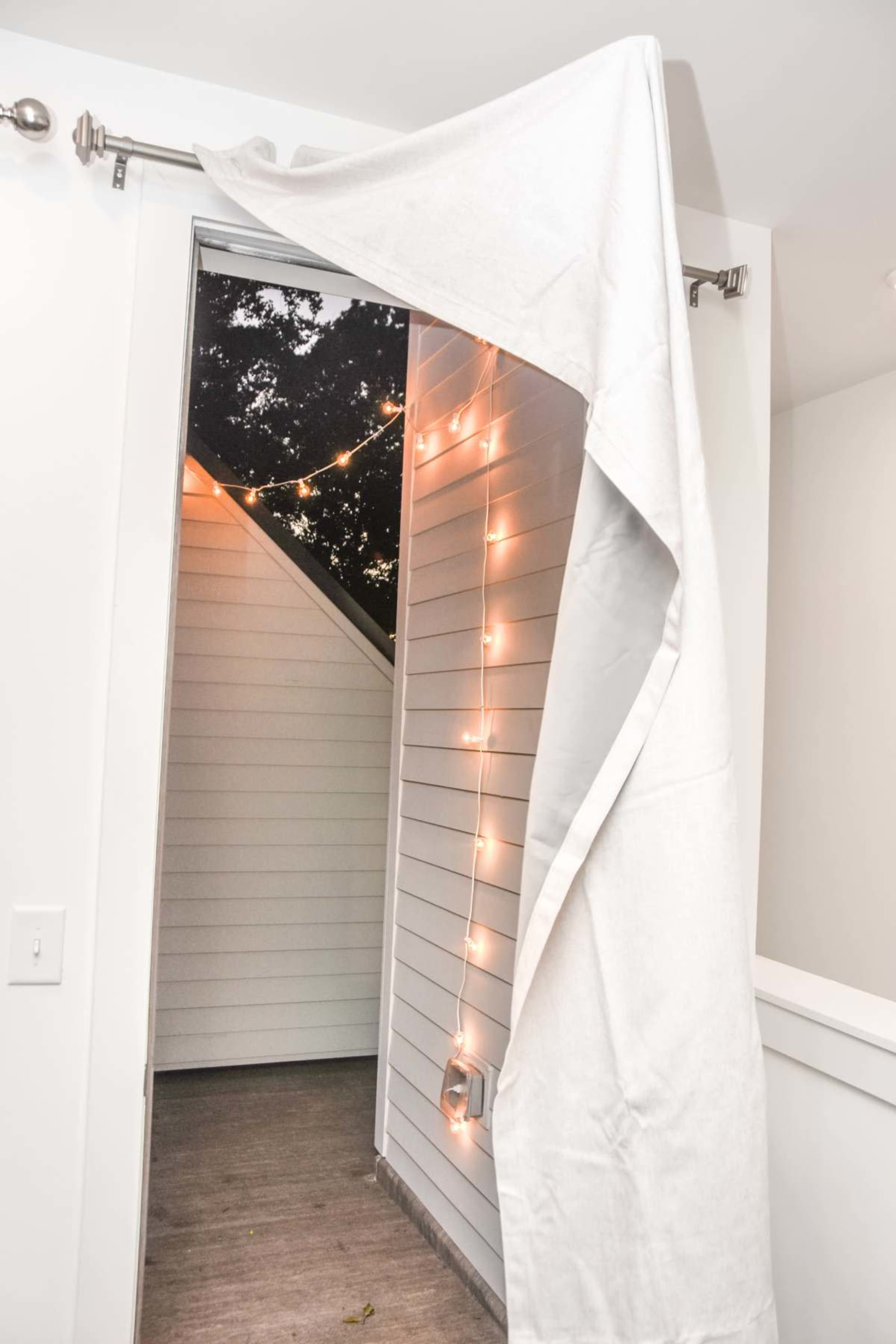 A white curtain is partially drawn back from an open doorway, revealing a small outdoor space adorned with string lights.