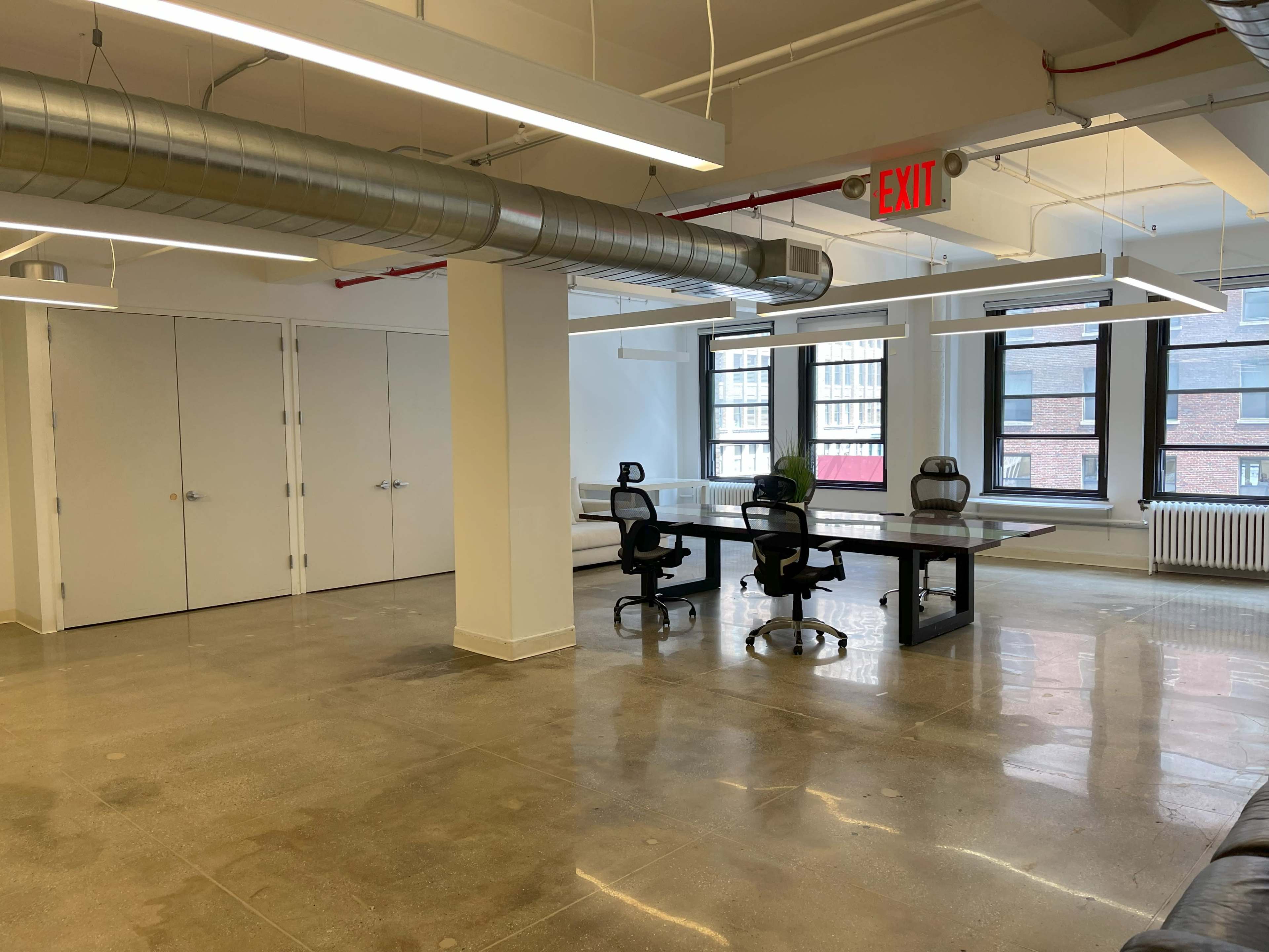 The image shows an empty office space with a large table and four chairs, featuring concrete floors and windows that allow natural light.