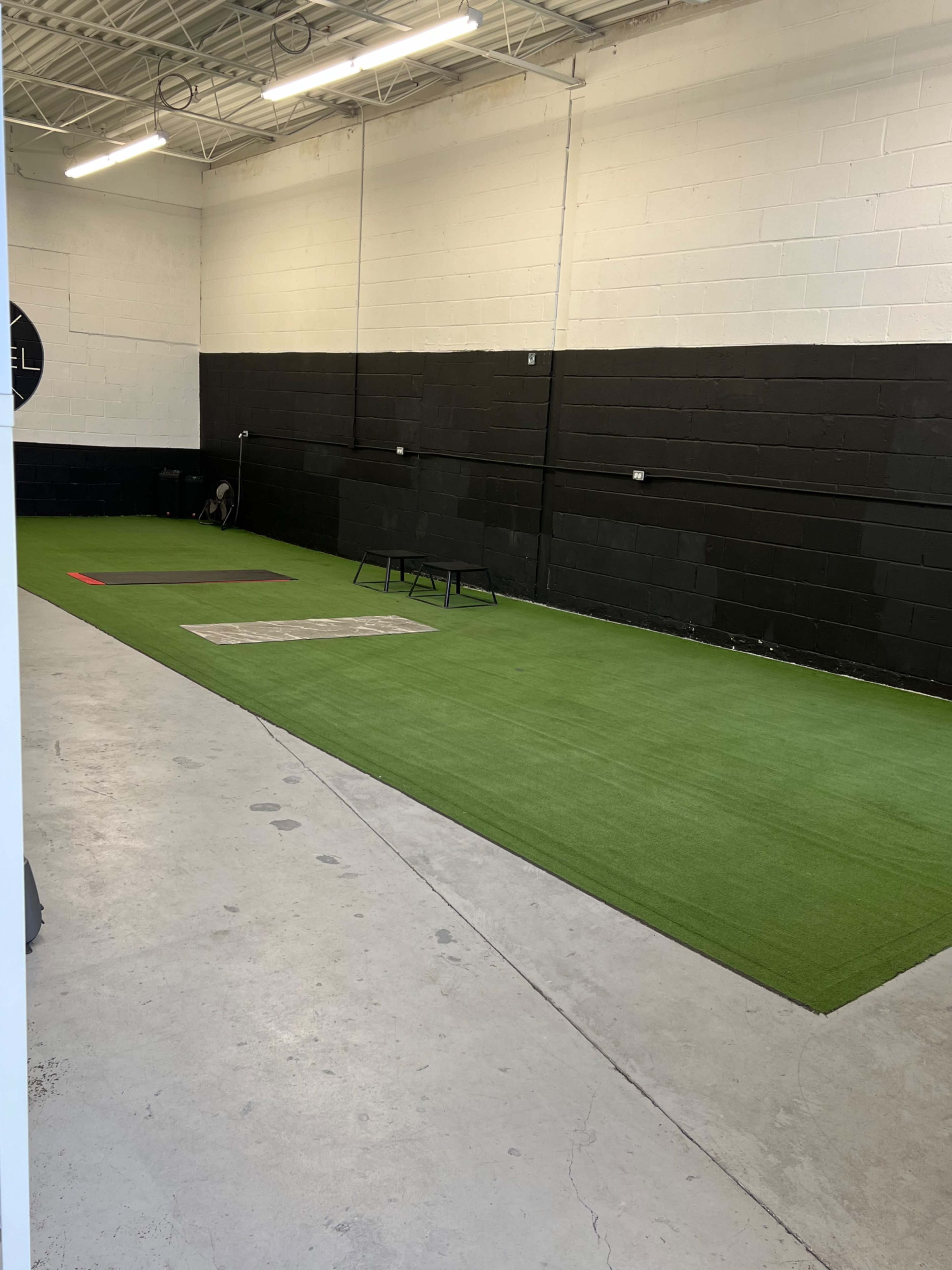 The image shows a spacious indoor area with artificial turf, marked training zones, and black walls.