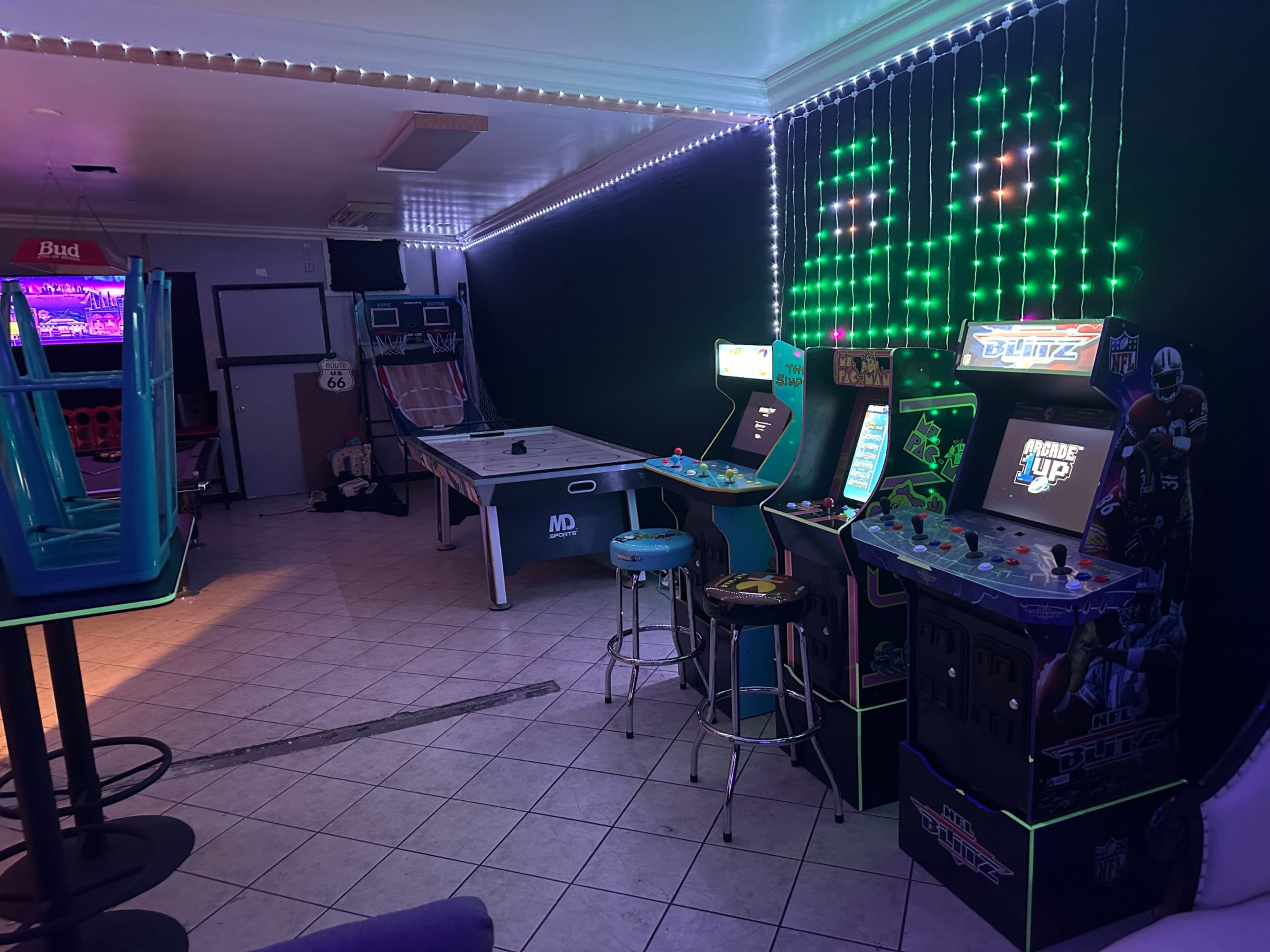 The image shows an arcade room featuring multiple gaming machines, including air hockey, retro arcade games, and neon lighting along the walls.