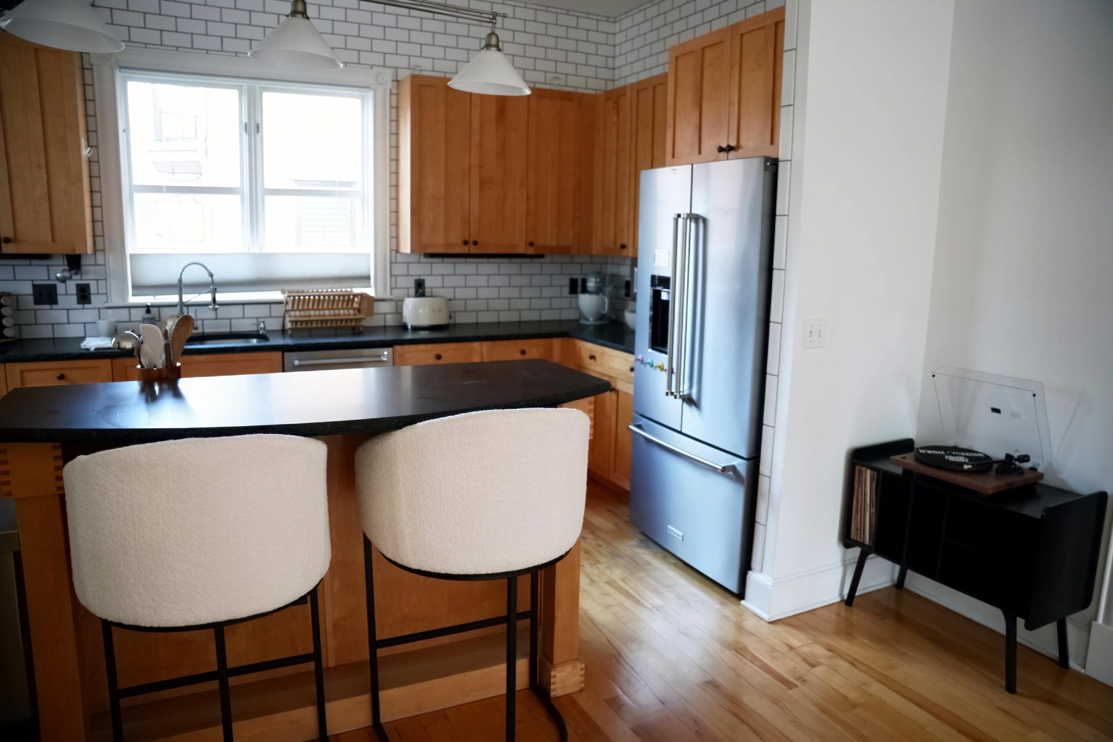 A modern kitchen with wooden cabinets, a black countertop, and a refrigerator, alongside a seating area with two bar stools and a vintage record player in the corner.