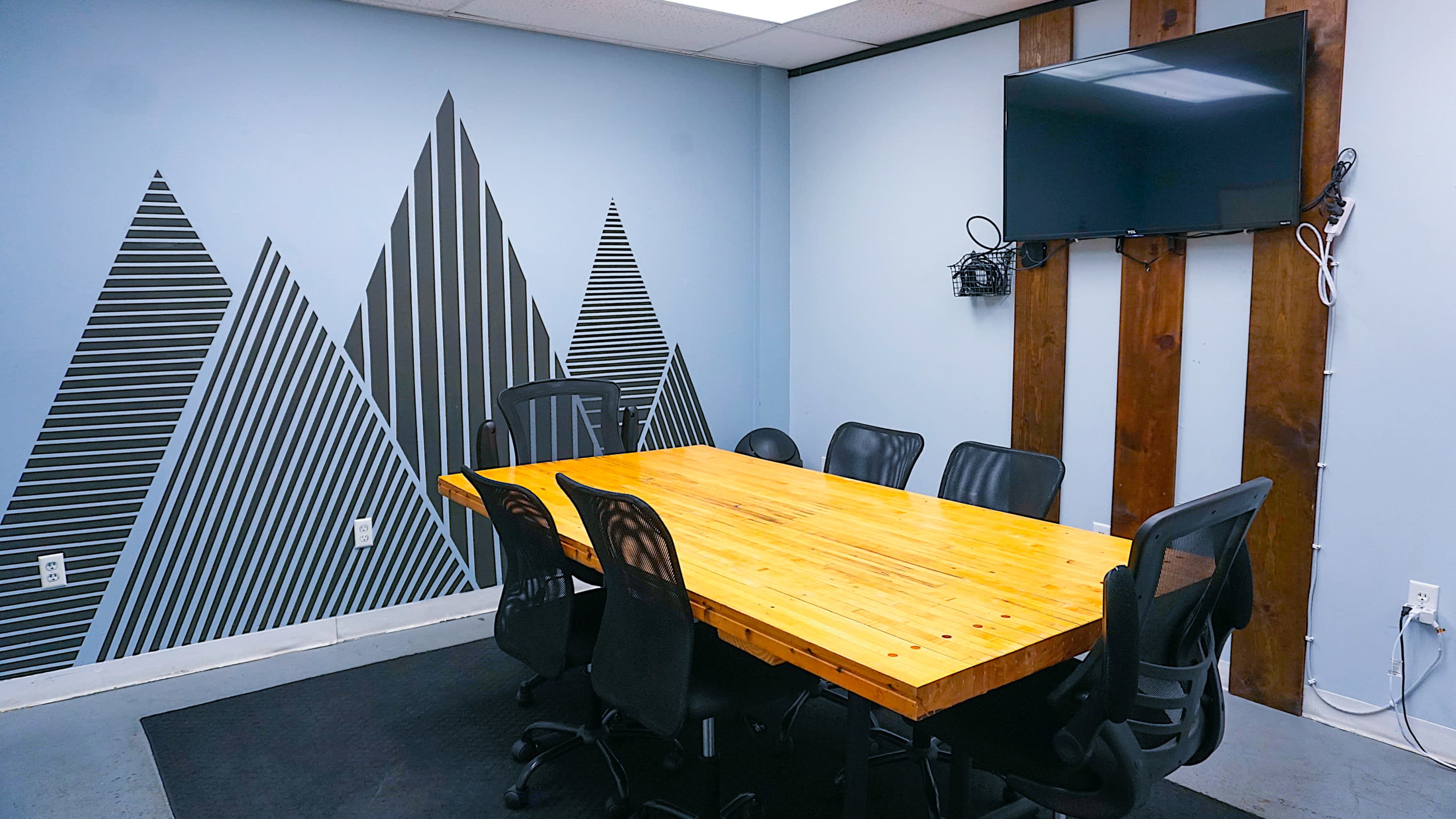 A conference room with a large wooden table surrounded by black chairs, featuring a wall decoration of striped mountains and a mounted television.