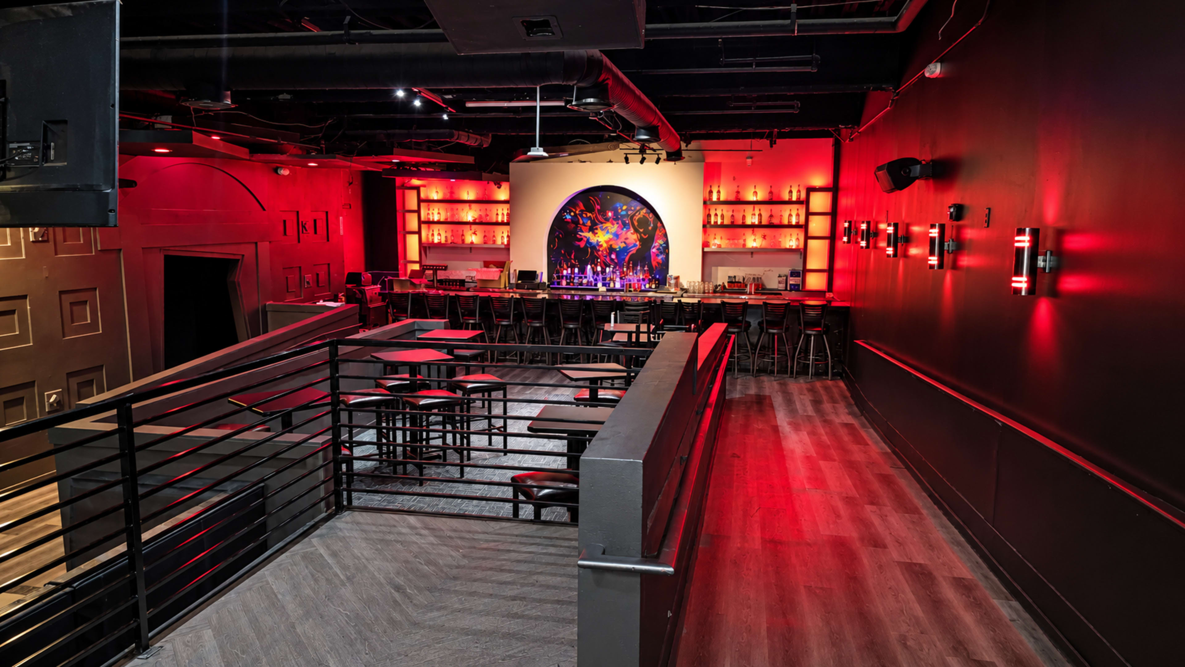 The image shows a modern bar setting with red lighting, featuring a long illuminated bar and elevated seating areas.