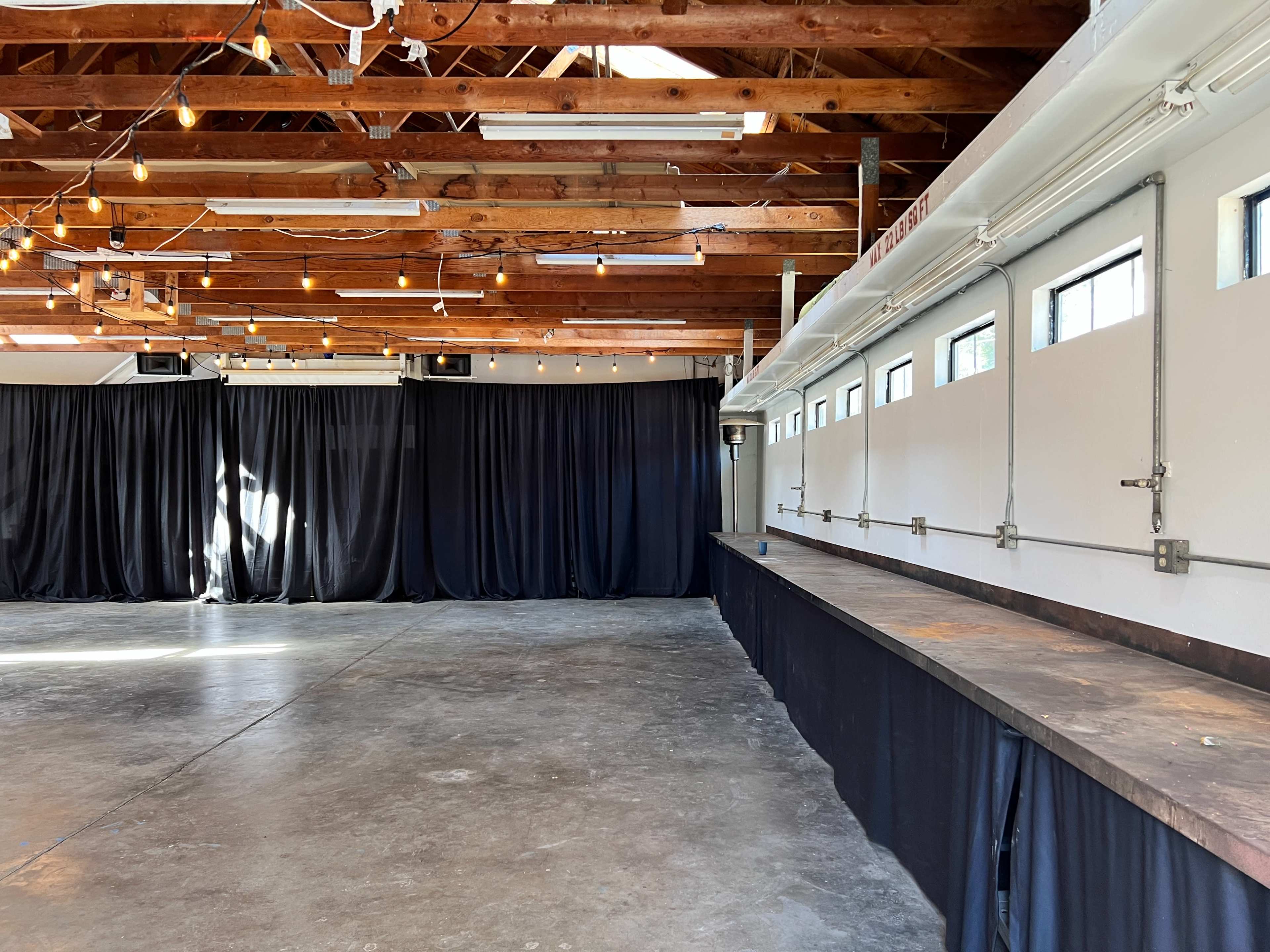 The image shows an indoor event space with wooden beams, string lights, a concrete floor, and a long counter next to a row of windows covered by black curtains.