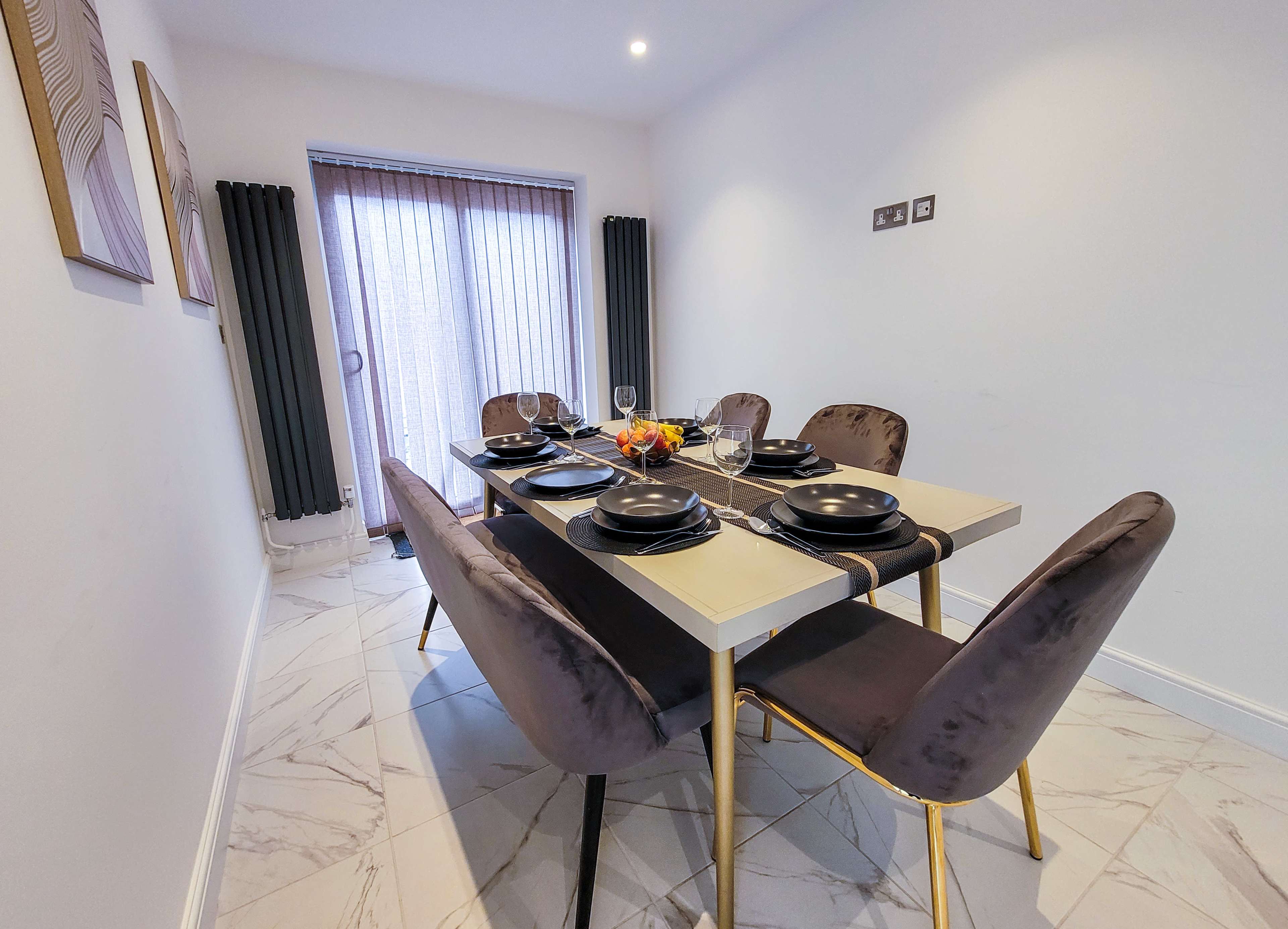 A dining table set for a meal with six black plates and wine glasses, surrounded by velvet chairs in a room with large windows.
