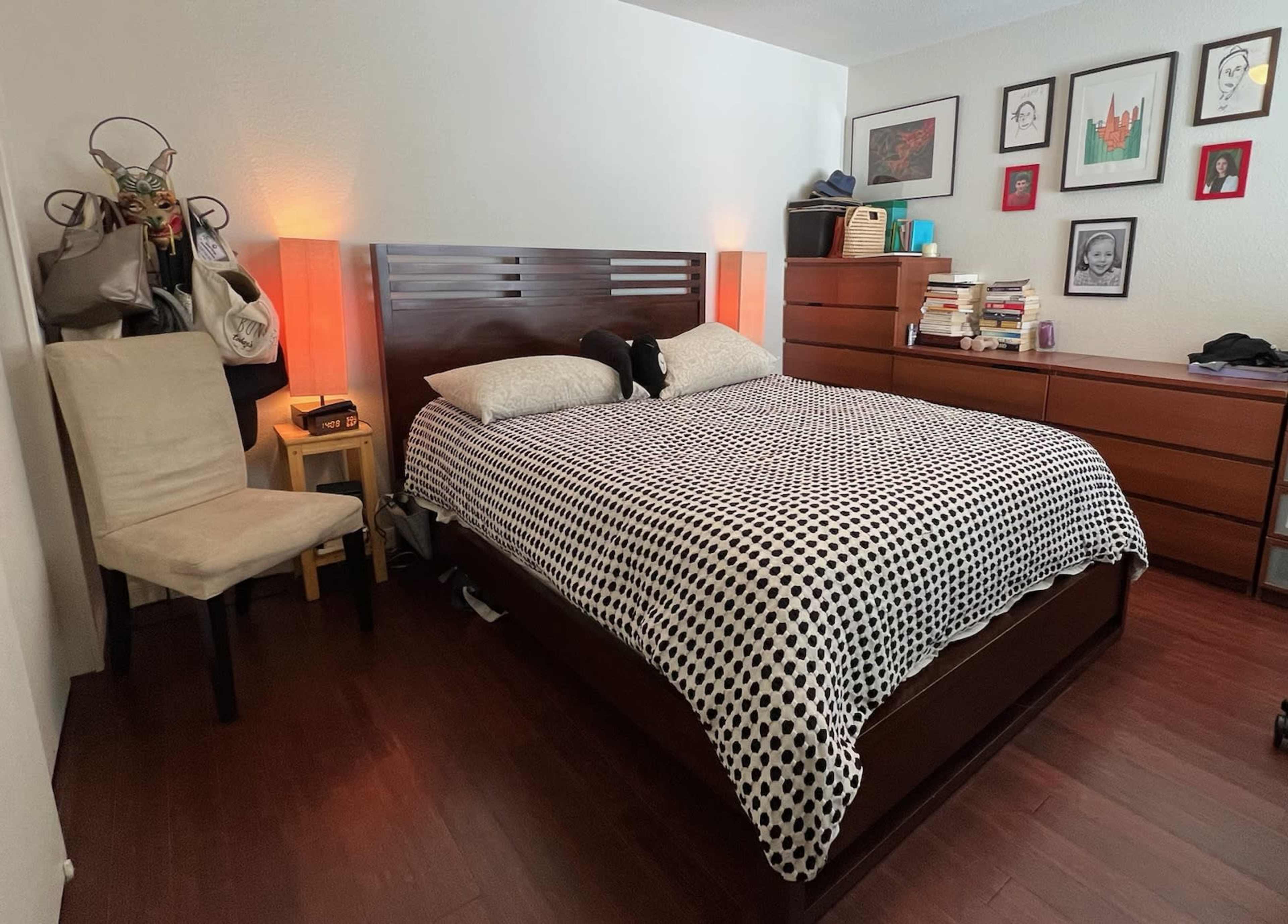 The image shows a neatly arranged bedroom featuring a bed with a patterned comforter, a chair, a wooden dresser, and framed artwork on the walls.