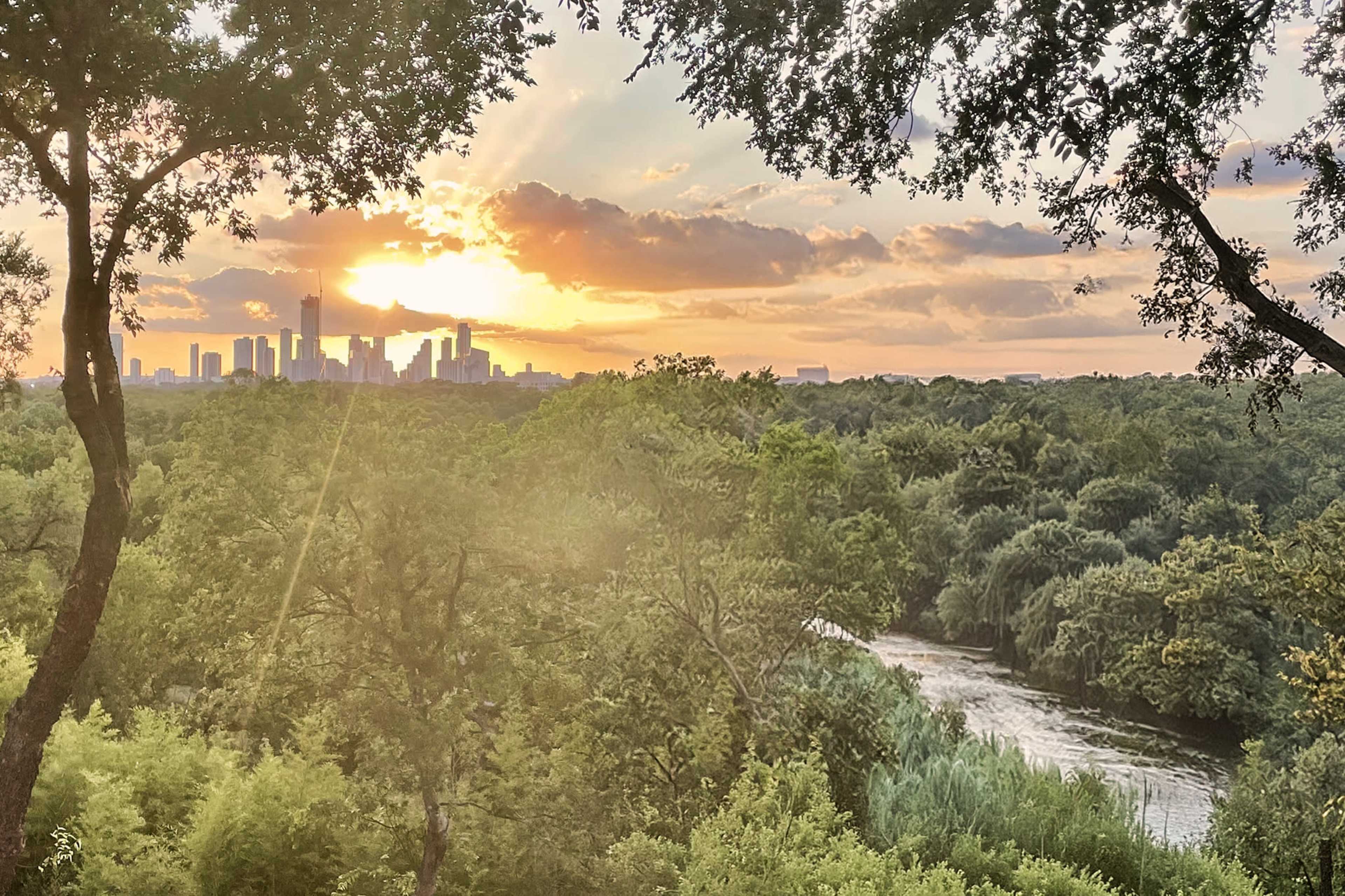 The sun sets behind a city skyline, casting light over a river bordered by dense greenery.