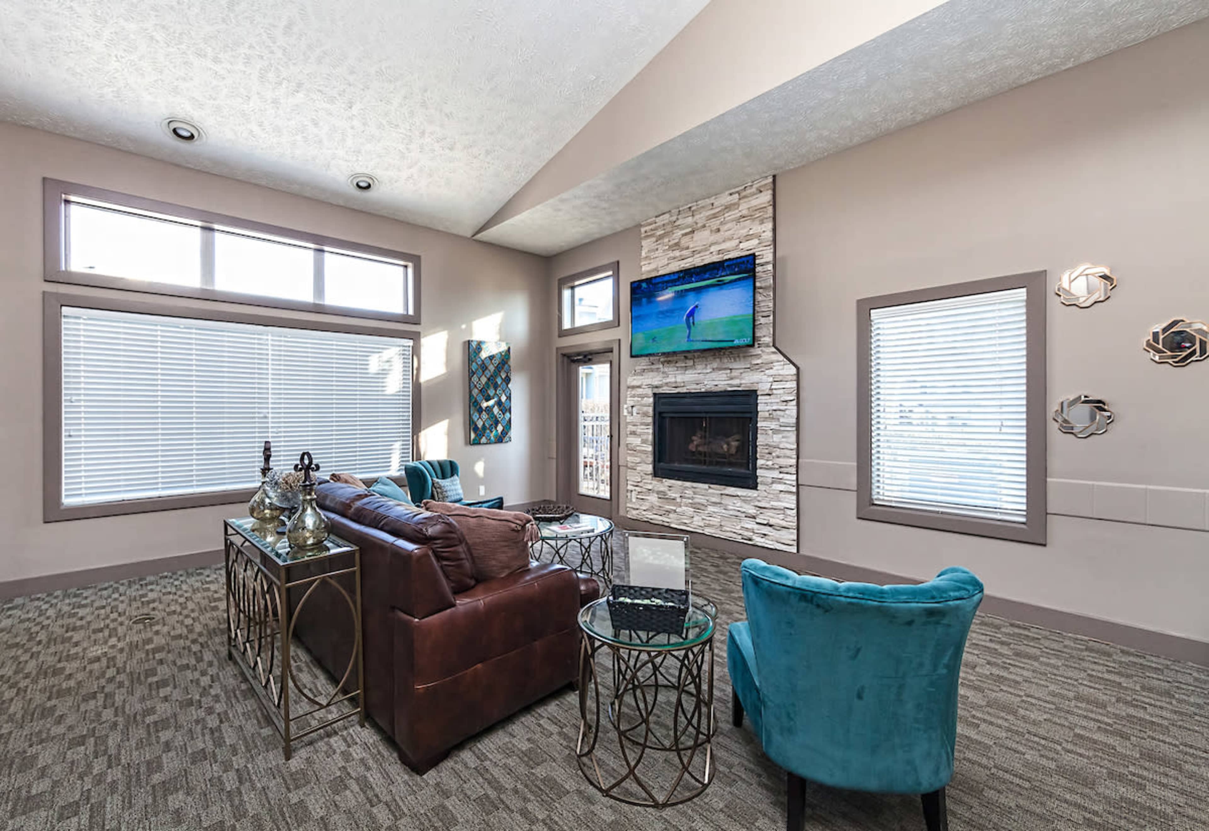 A modern living room with a brown leather sofa, teal accent chairs, a stone fireplace, and large windows allowing natural light to enter.