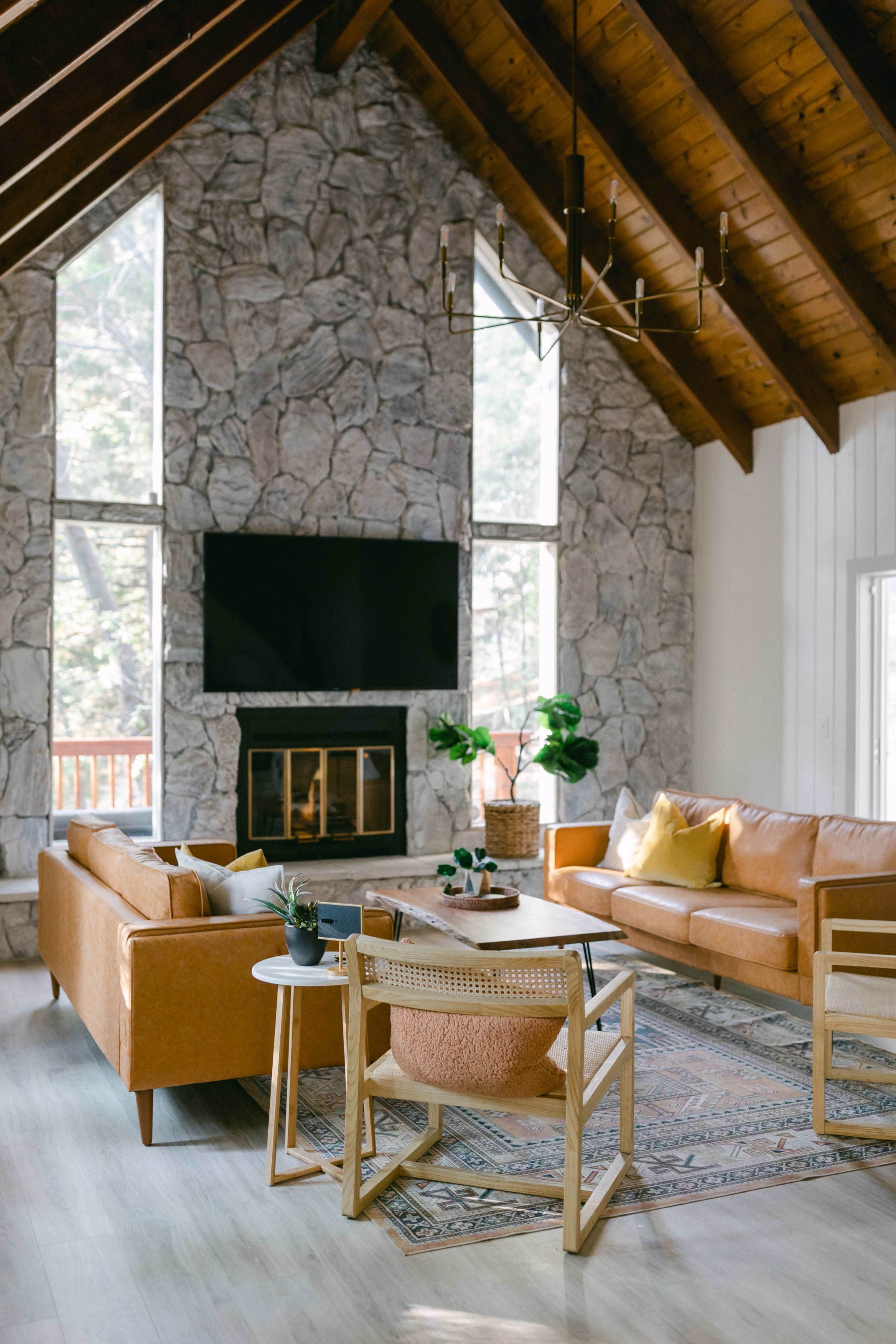 A spacious living room features a high, wooden-beamed ceiling, stone walls, a mounted TV, two brown leather sofas, and a light-colored area rug.
